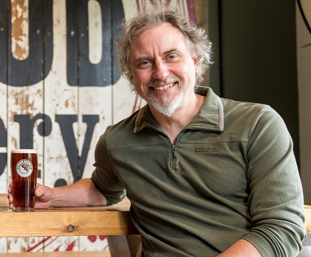 A man wearing a green fleece is sitting with a pint of beer in his hand. The pint glass says Stroud Bewery.