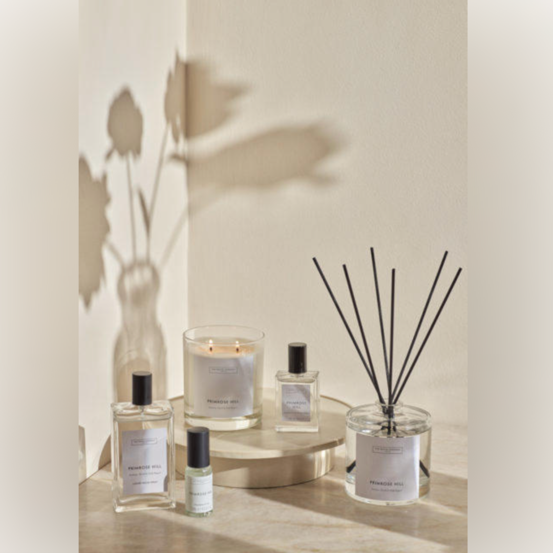 A reed diffuser, a candle and perfume bottles are photographed in front of a cream wall