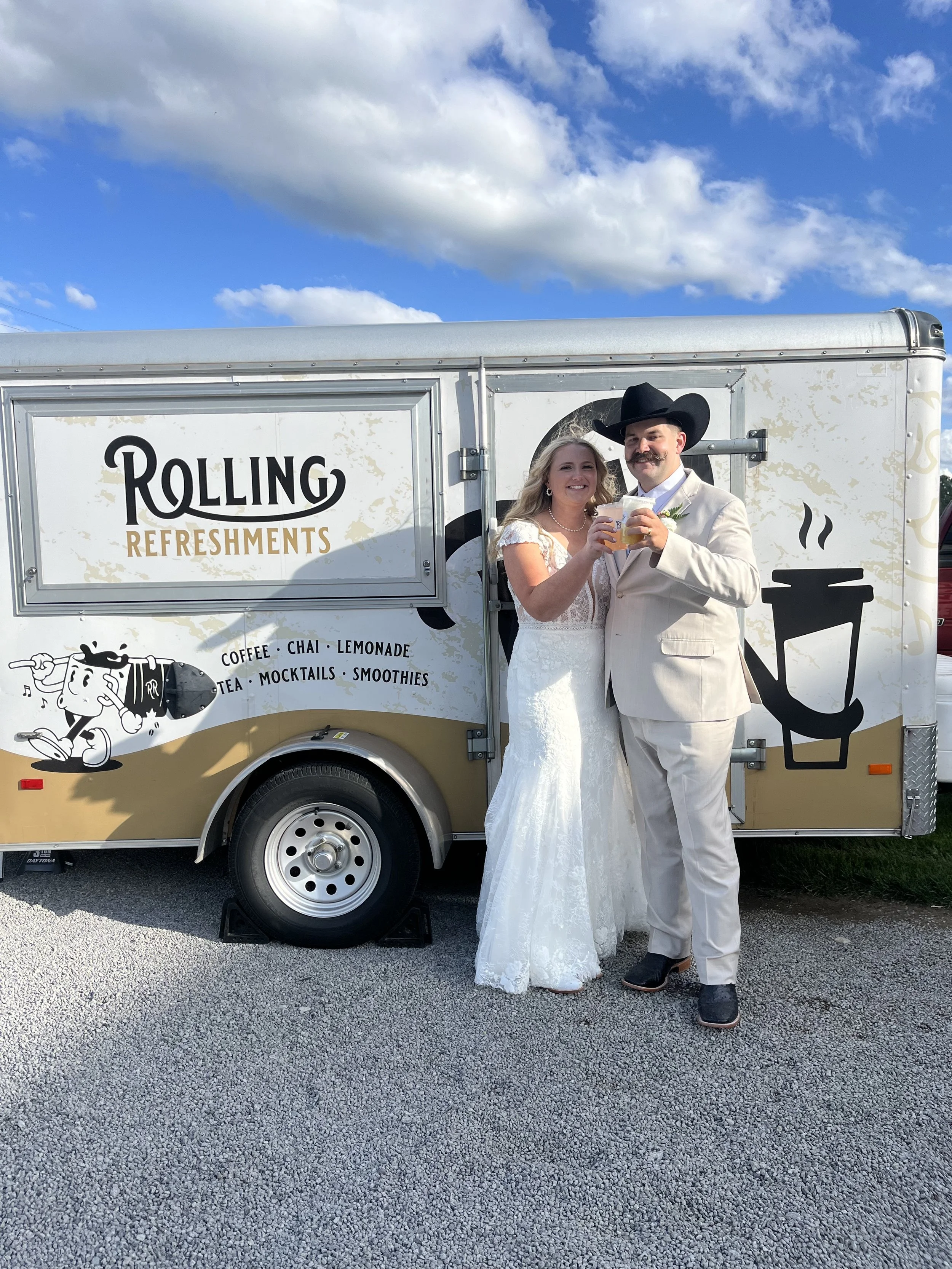 A bride and groom wearing wedding attire are standing in front of a food truck, smiling and holding drinks together against a cloudy sky.