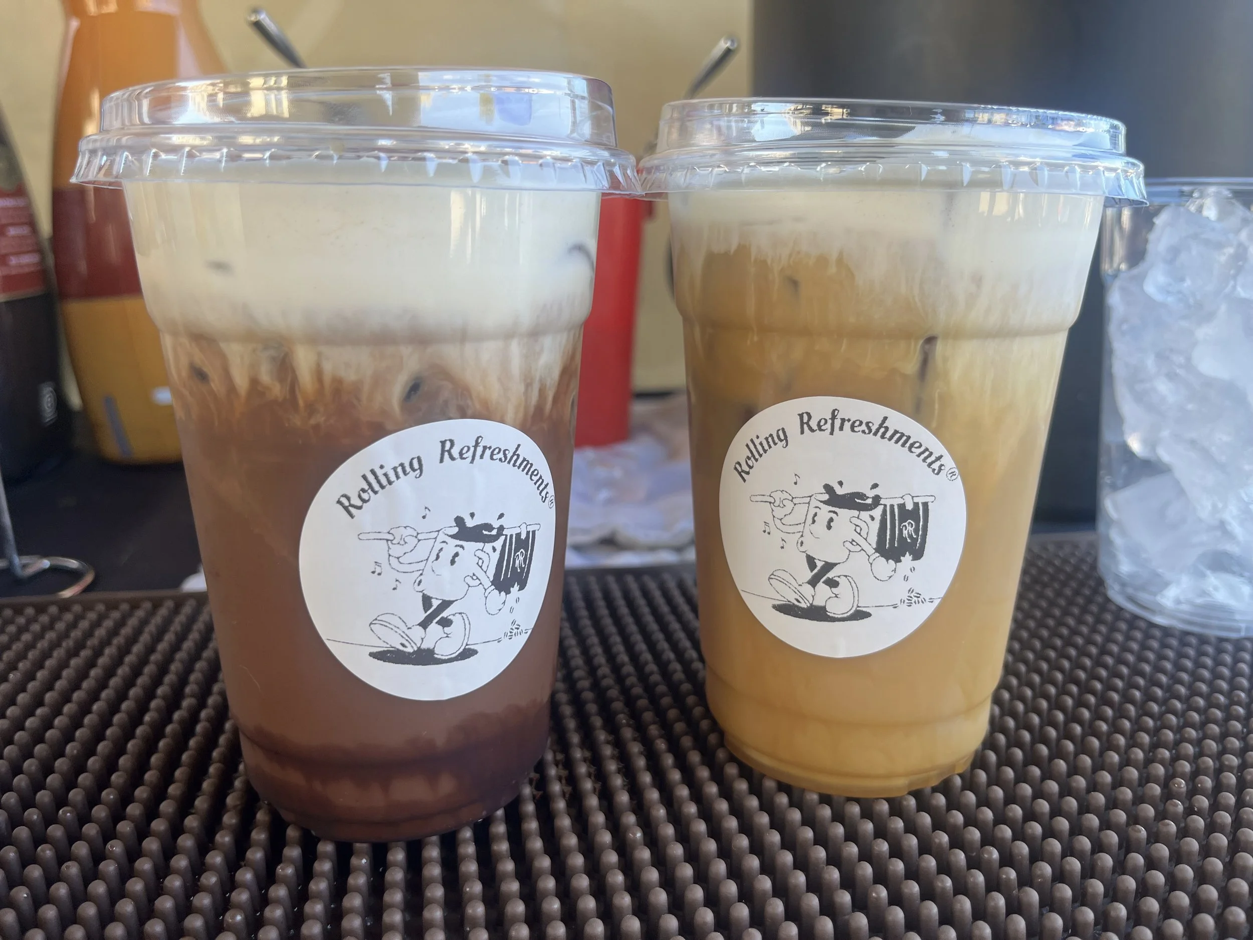 Two iced coffee drinks in clear plastic cups with whipped cream on top, branded with a sticker that says "Rolling Refreshments" and features a cartoon character holding a tray with a beverage.
