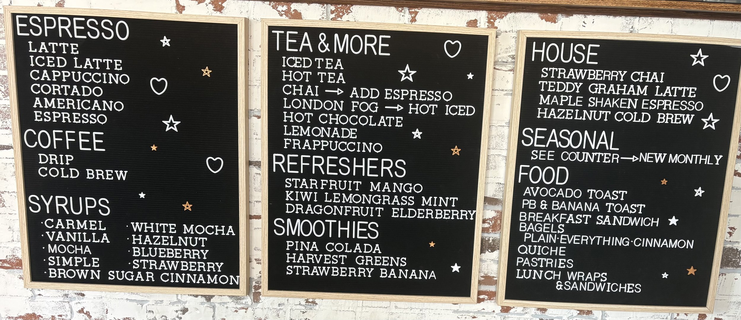 Three black menu boards with white text and orange and white star and heart decorations. The left board lists espresso drinks, coffee, and syrups; the middle board lists teas, refreshments, and smoothies; the right board lists house specialties, seasonal items, and food options.