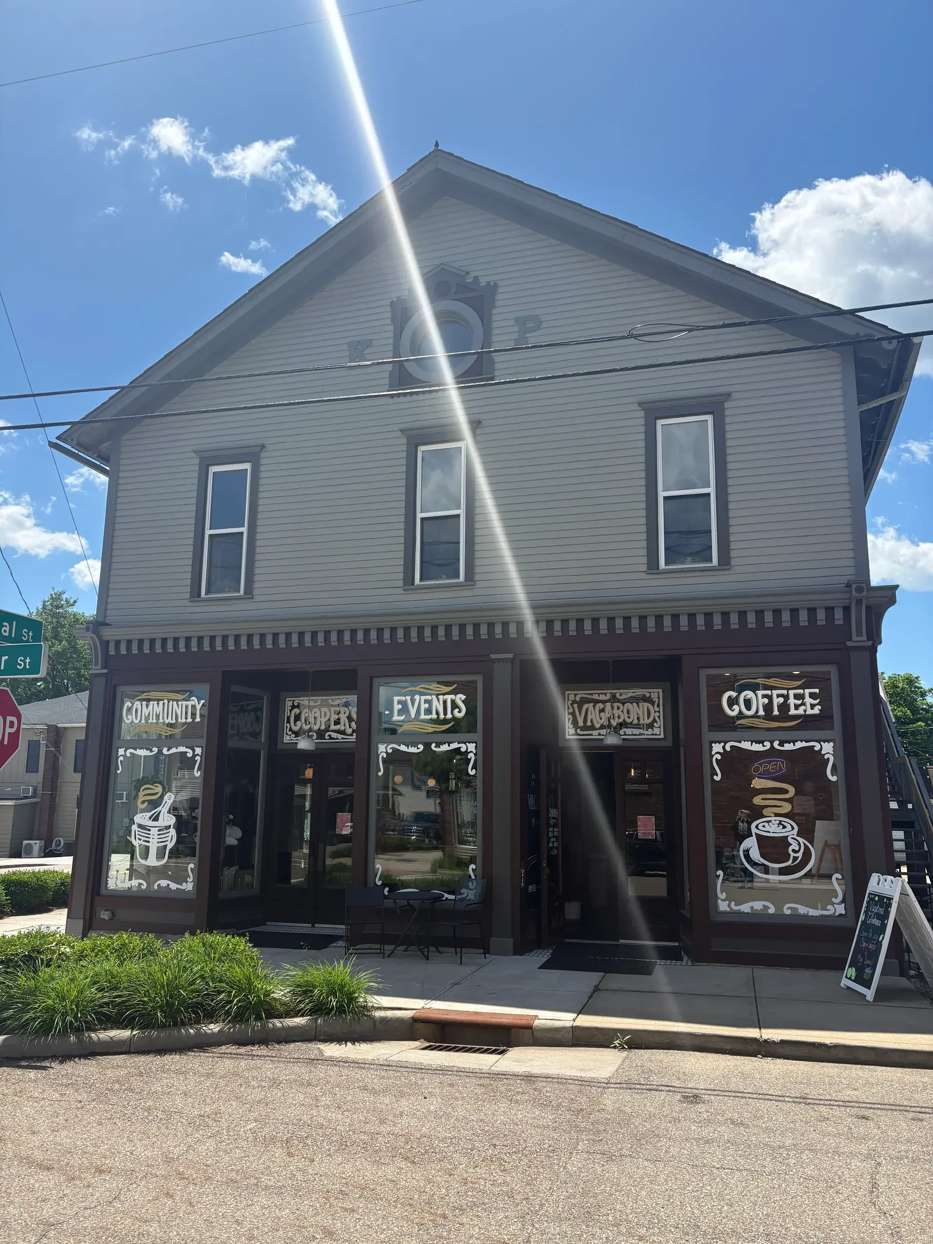 A two-story building with a coffee shop on the ground floor, featuring large windows with coffee-themed graphics, and a sign indicating it offers community events and is open for business. The building's upper part has light-colored siding, multiple tall windows, and a decorative clock face. The sky is blue with a few clouds, and a bright sunlight beam creates a lens flare in the image.