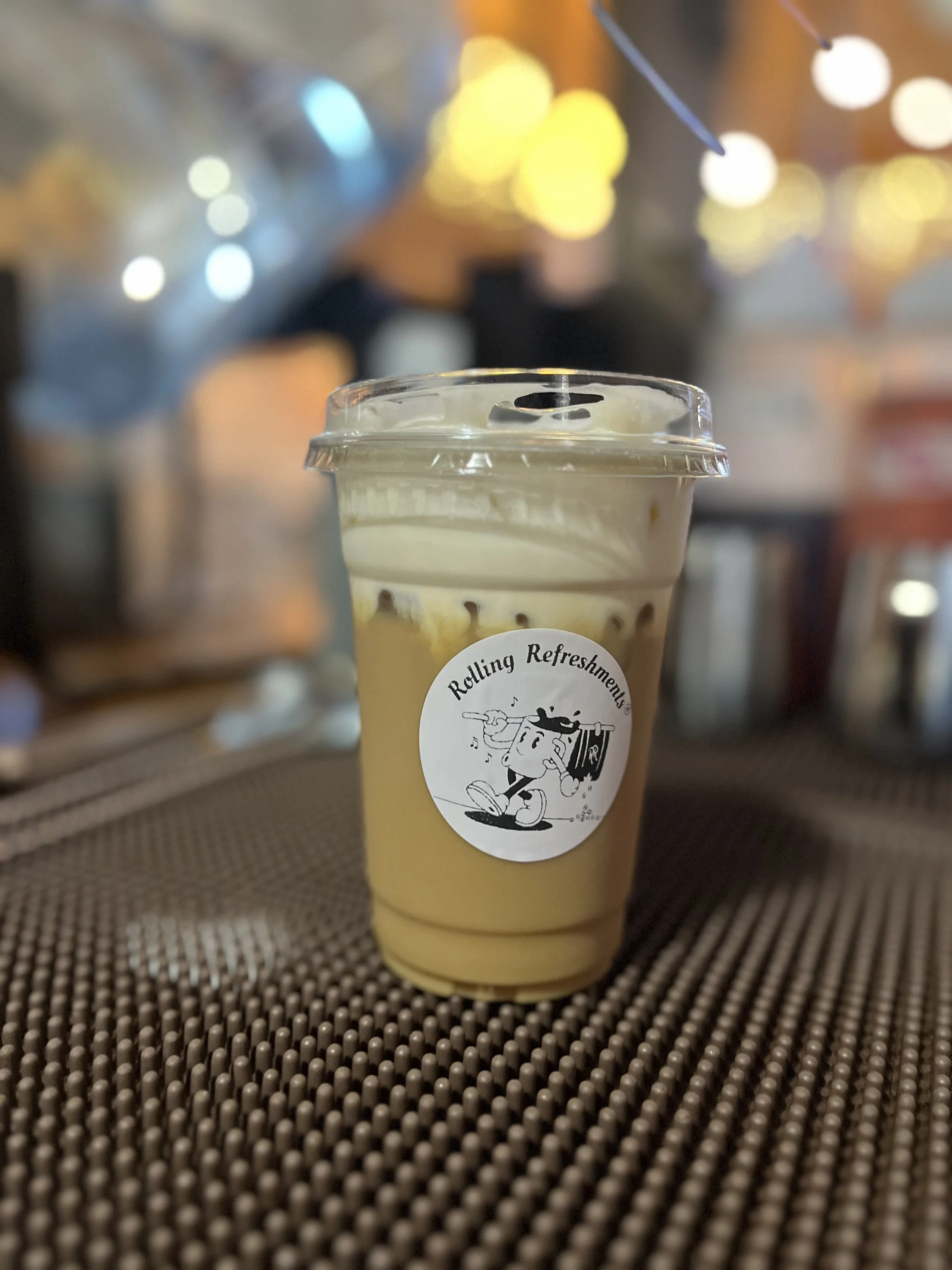 Plastic cup with a sealed lid containing an iced beverage, sitting on a textured brown placemat with blurred colorful lights in the background, and a sticker that says "Rolling Refreshments" with a cartoon character playing an electric guitar.