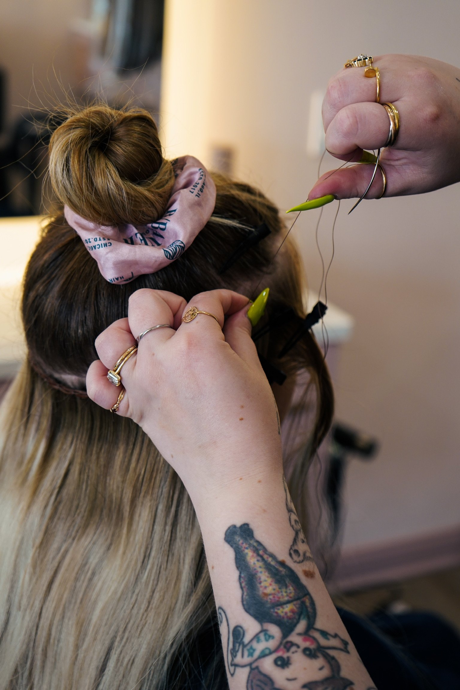 A person with long blonde hair getting an eyelash extension, with the eyelash technician using tools and wearing multiple rings, including a gold ring with a large gemstone and a silver ring. The person has a tattoo of a bear and a cartoon girl on their arm.