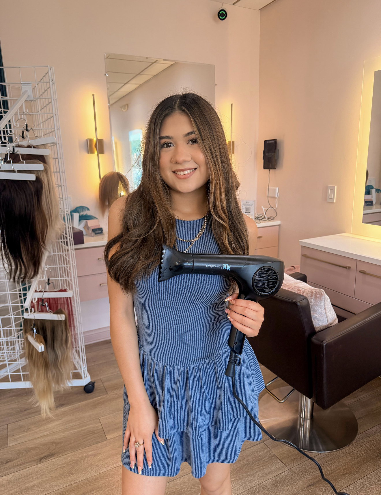 A young woman with long brown hair, wearing a blue dress, holds a black hairdryer in a salon with pink and white walls, mirrors, and wigs.