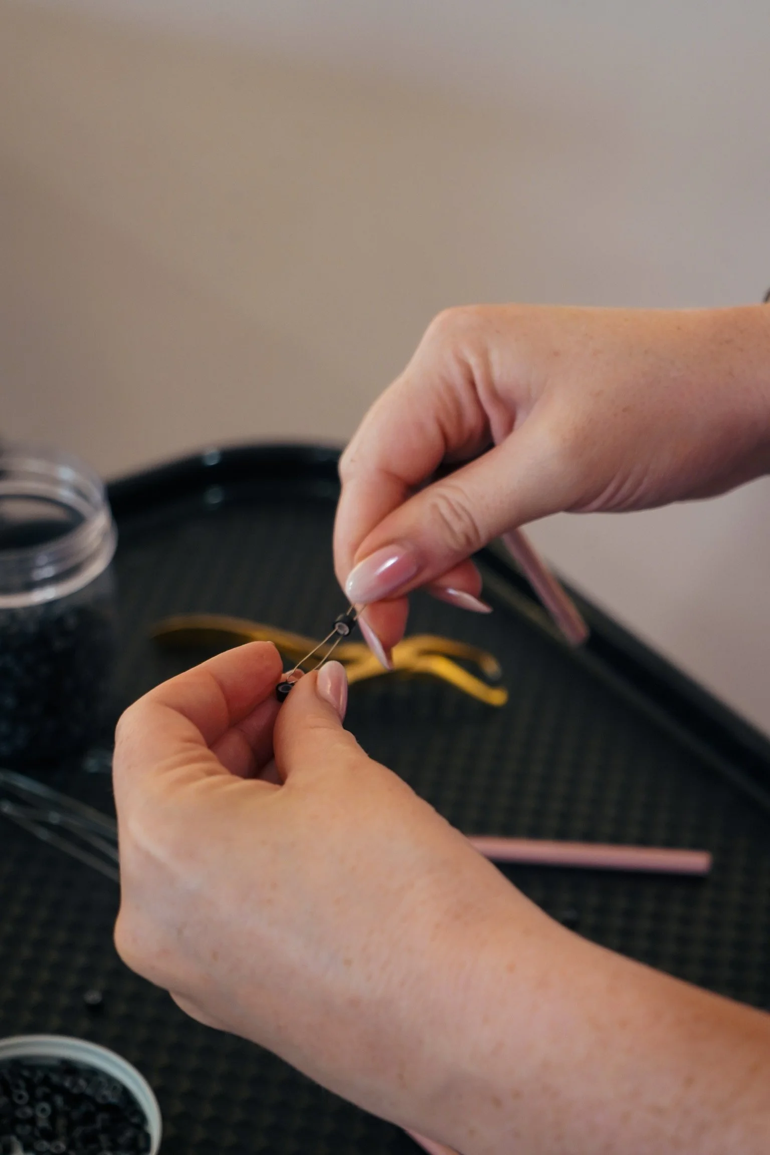Close-up of a person's hands threading a tiny needle with black beads, with various beading supplies on a black tray.