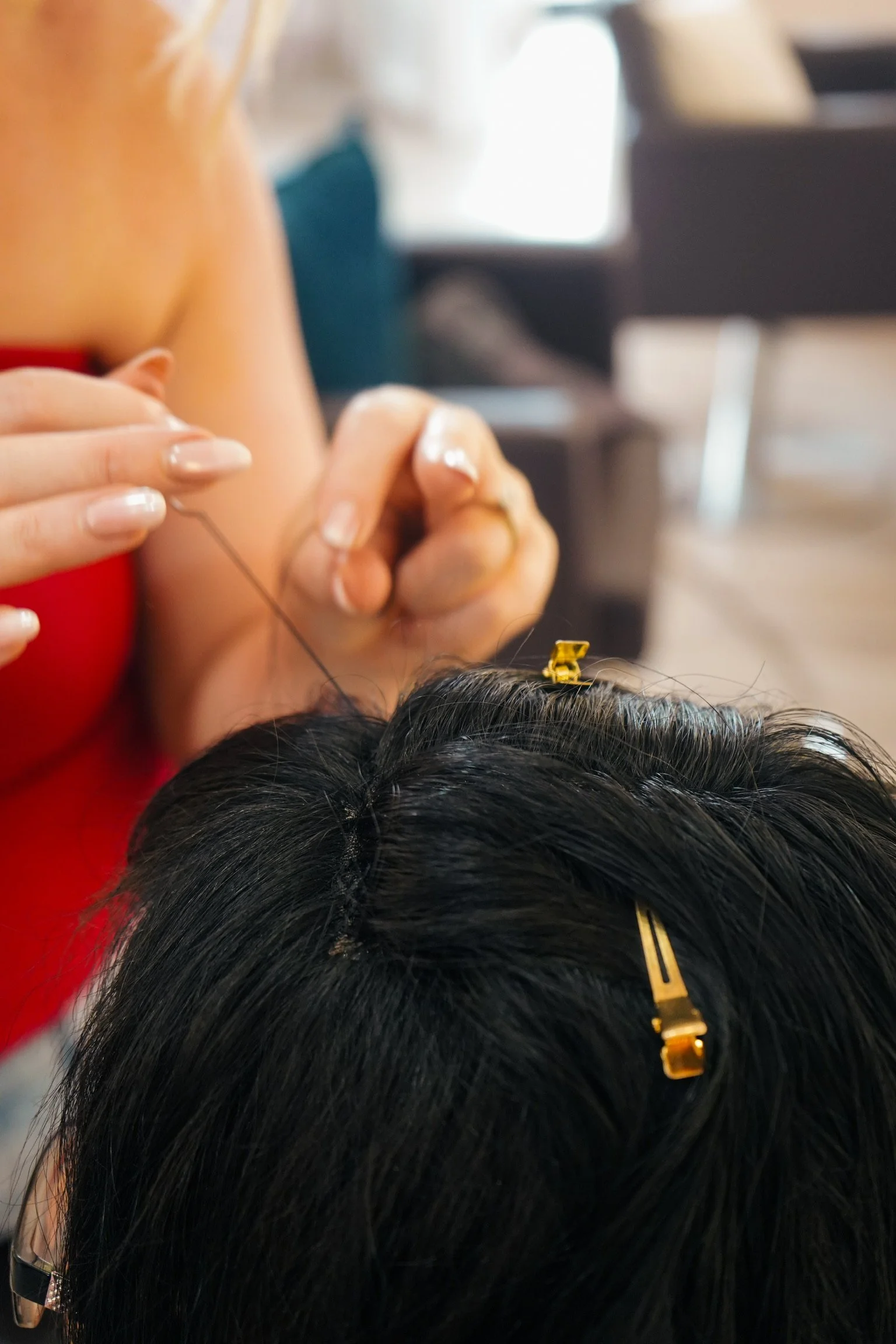 A woman with long dark hair getting a hair weave installed. Hair clips hold her hair back, and a hairstylist is using a needle and thread to sew the weave into her scalp.