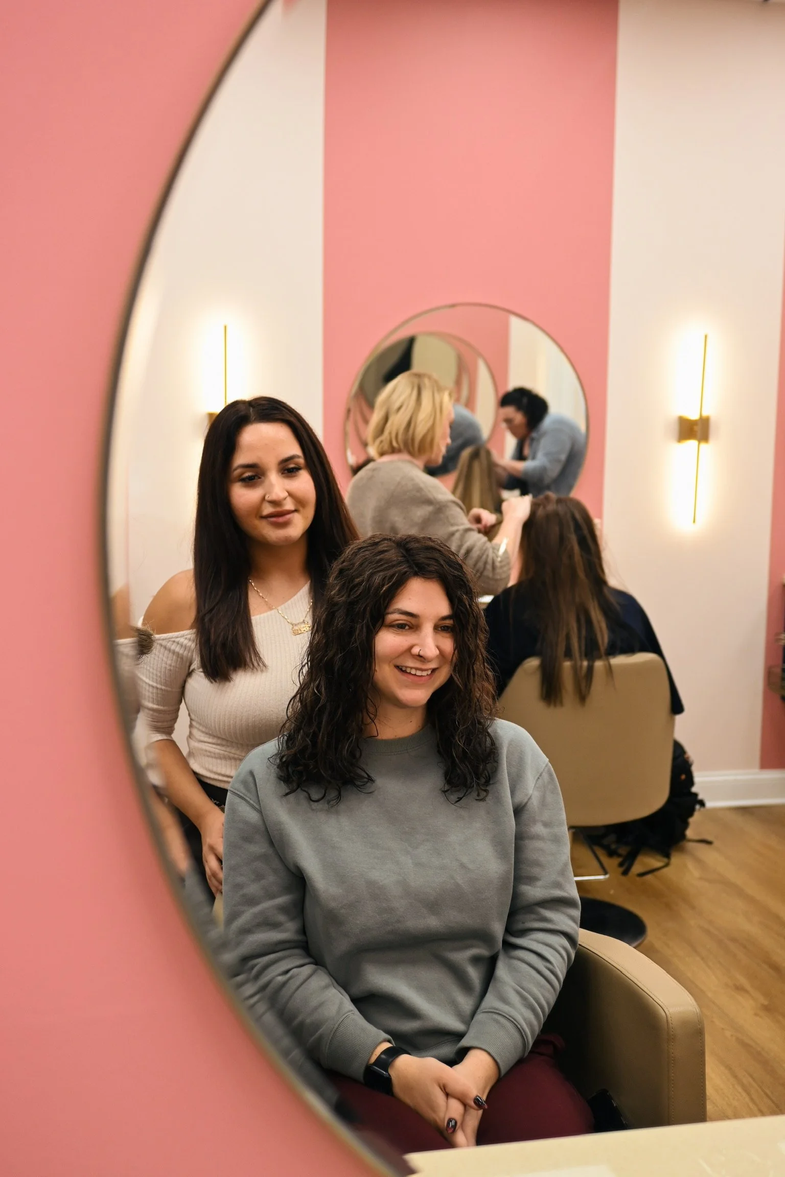 Two women sitting in a salon, with one in the mirror and the other behind her, getting hair styled.