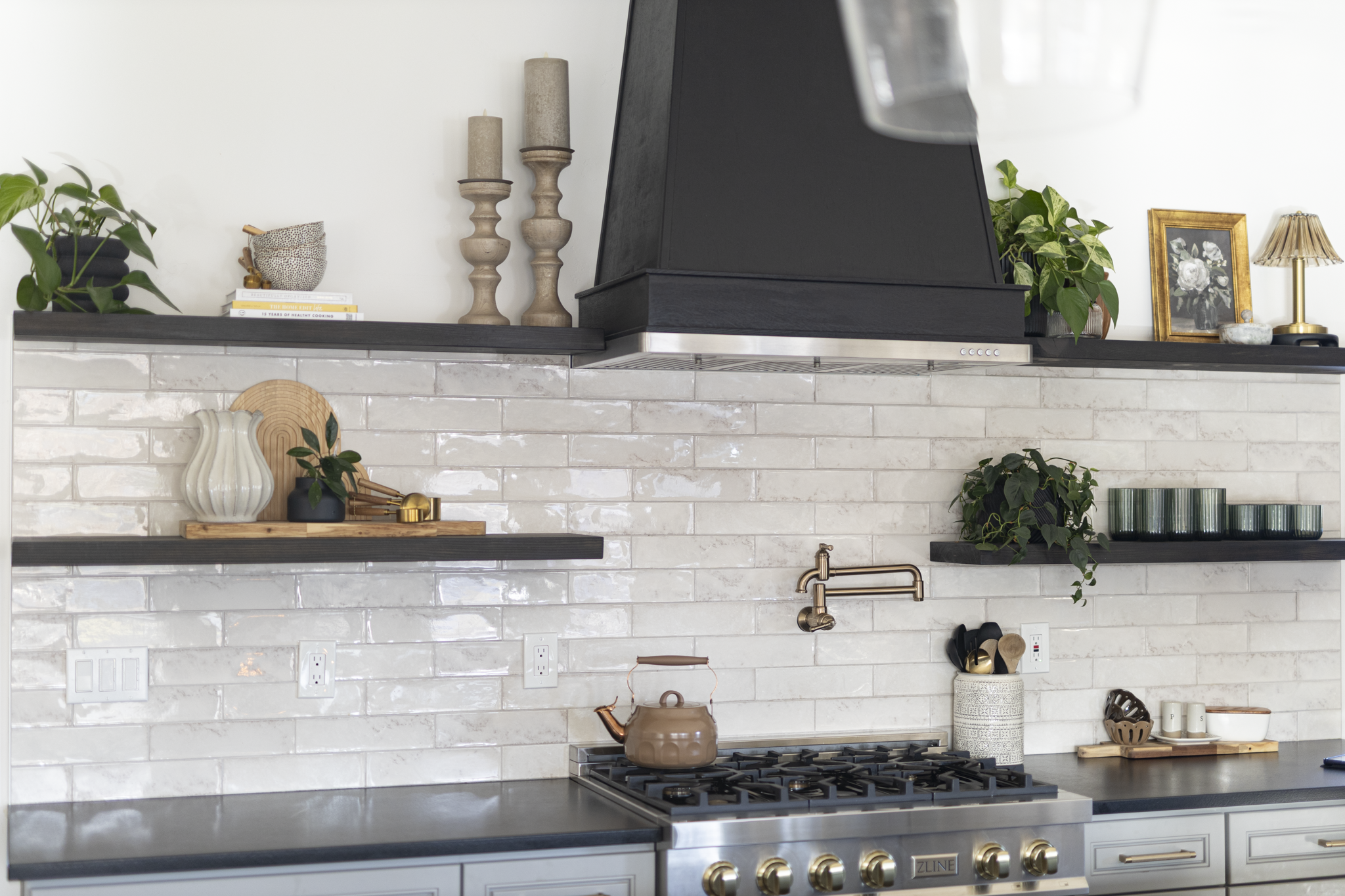Modern kitchen with beige brick backsplash, black open shelves decorated with plants and decorative items, a stainless steel stove with a brown teapot, and a black range hood.