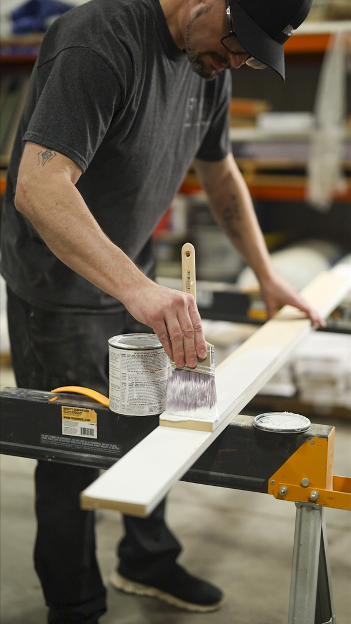 A man in a black t-shirt and glasses is painting a piece of wood white in a workshop, using a paintbrush from a can of white paint.