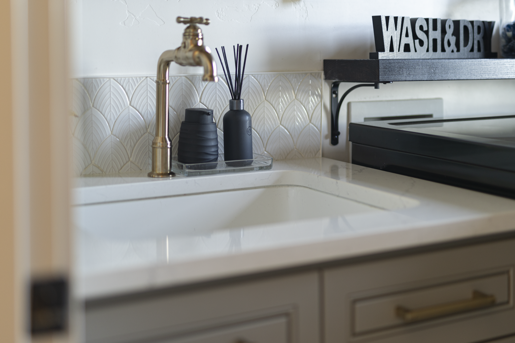 Kitchen sink with a gold-colored faucet, black soap dispenser, black diffuser, and a clear tray, with a black shelf above holding a black sign that says 'WASH & DRY'.