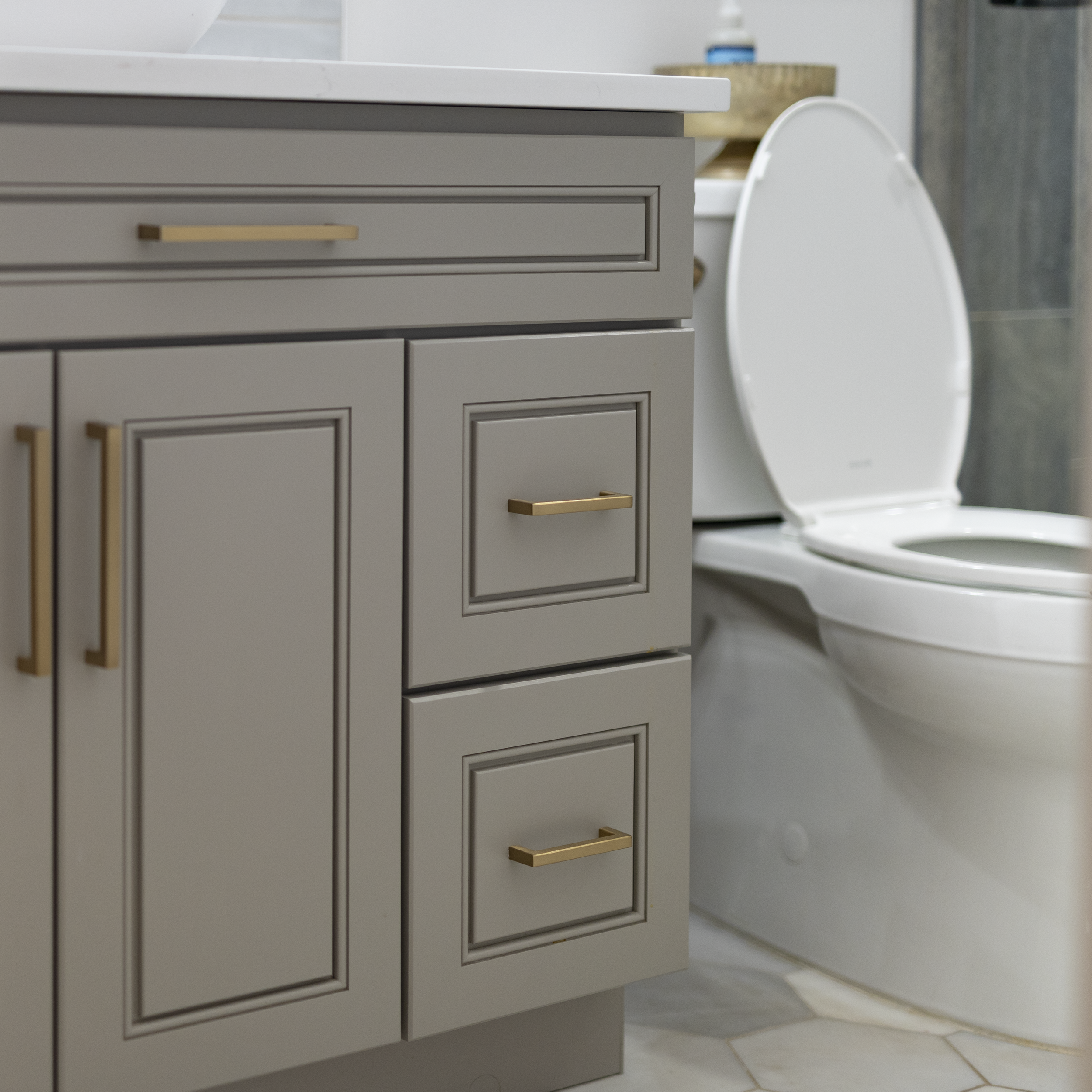 Close-up of a gray bathroom vanity with gold handles and a white toilet in the background.