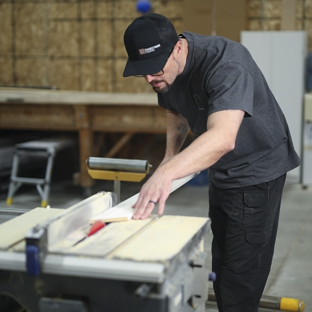 Man wearing a black cap and glasses working with a table saw in a woodworking shop
