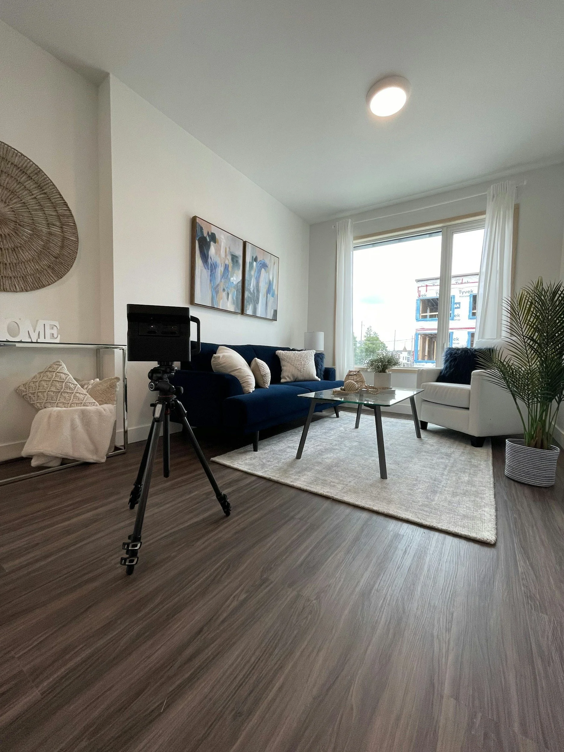 Modern living room with a blue sofa, coffee table, and neutral decor. Large windows provide natural light. A tripod is set up on the wooden floor. An indoor plant is placed near an armchair.