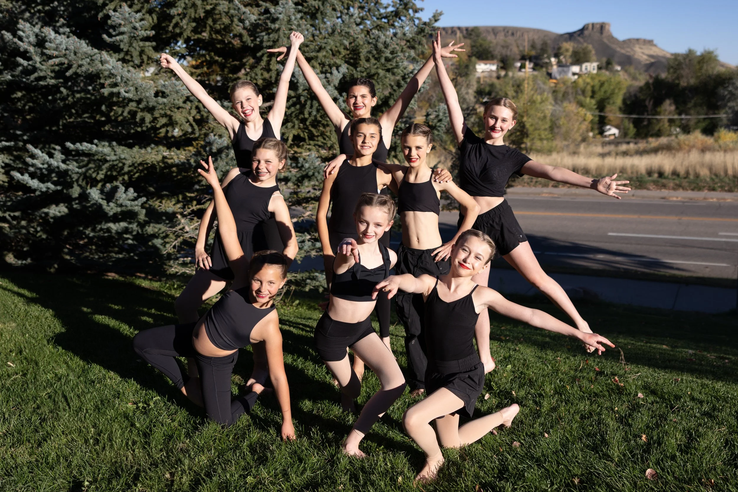 A group of twelve young female dancers in black dance outfits posing outdoors in front of a large pine tree and a backdrop of autumn trees and mountains.
