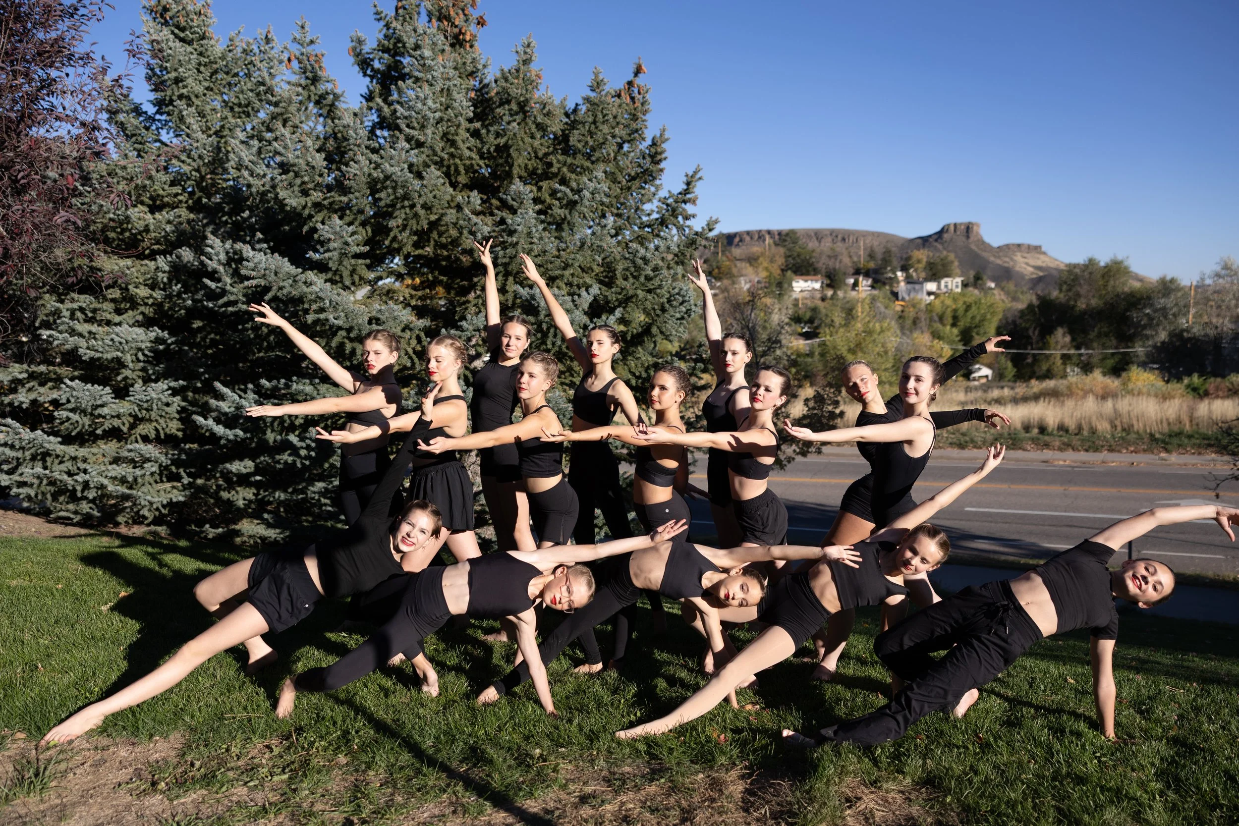 A group of seven young female dancers in black dance attire posing outdoors on a grassy area with tall evergreen trees and fall foliage in the background.