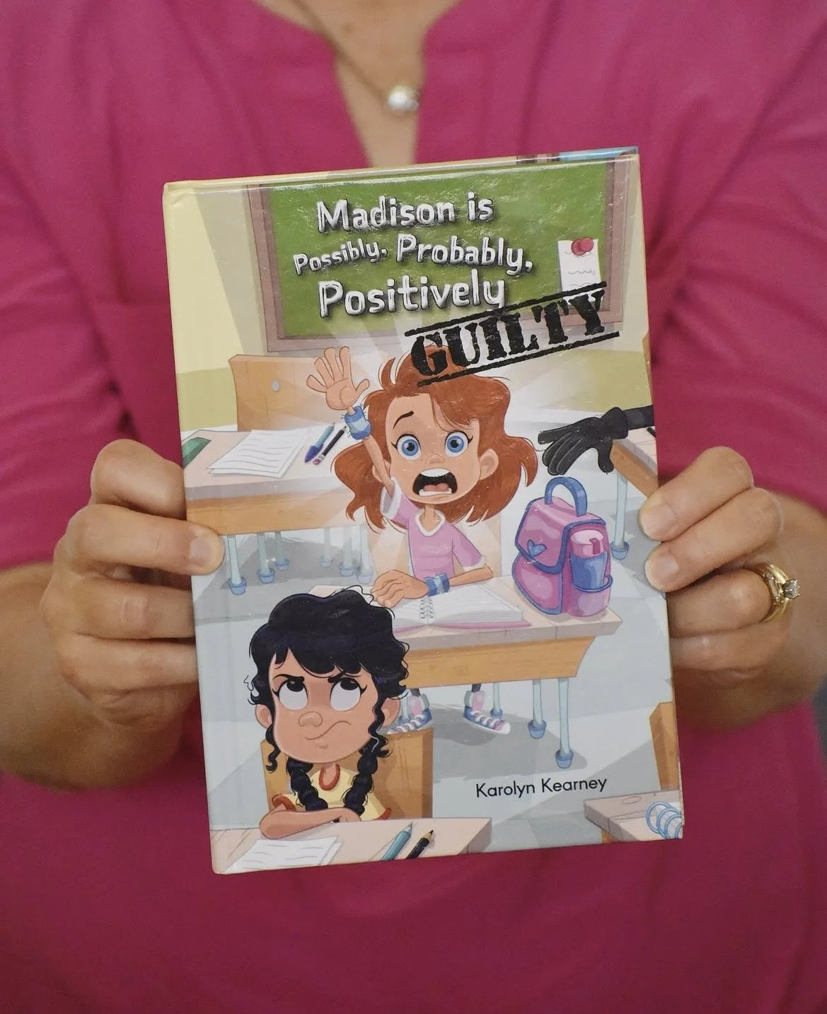 Close-up of hands holding the children’s book Madison is Possibly, Probably, Positively Guilty with a bright pink background