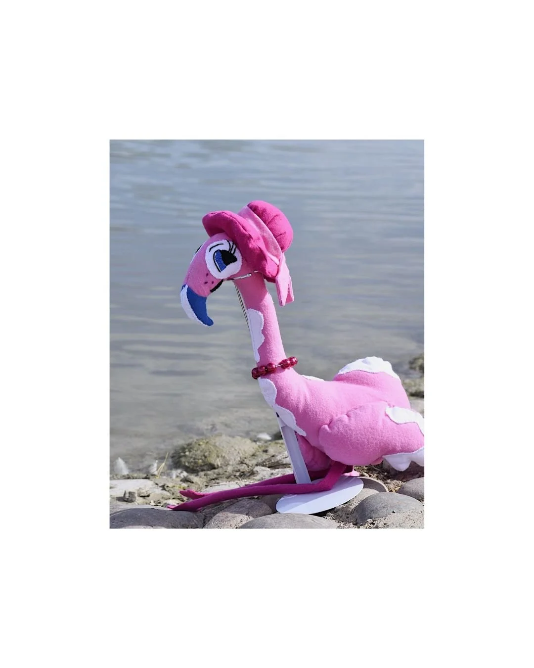 Pink flamingo plush toy placed by the water on rocks in an outdoor lifestyle product photo