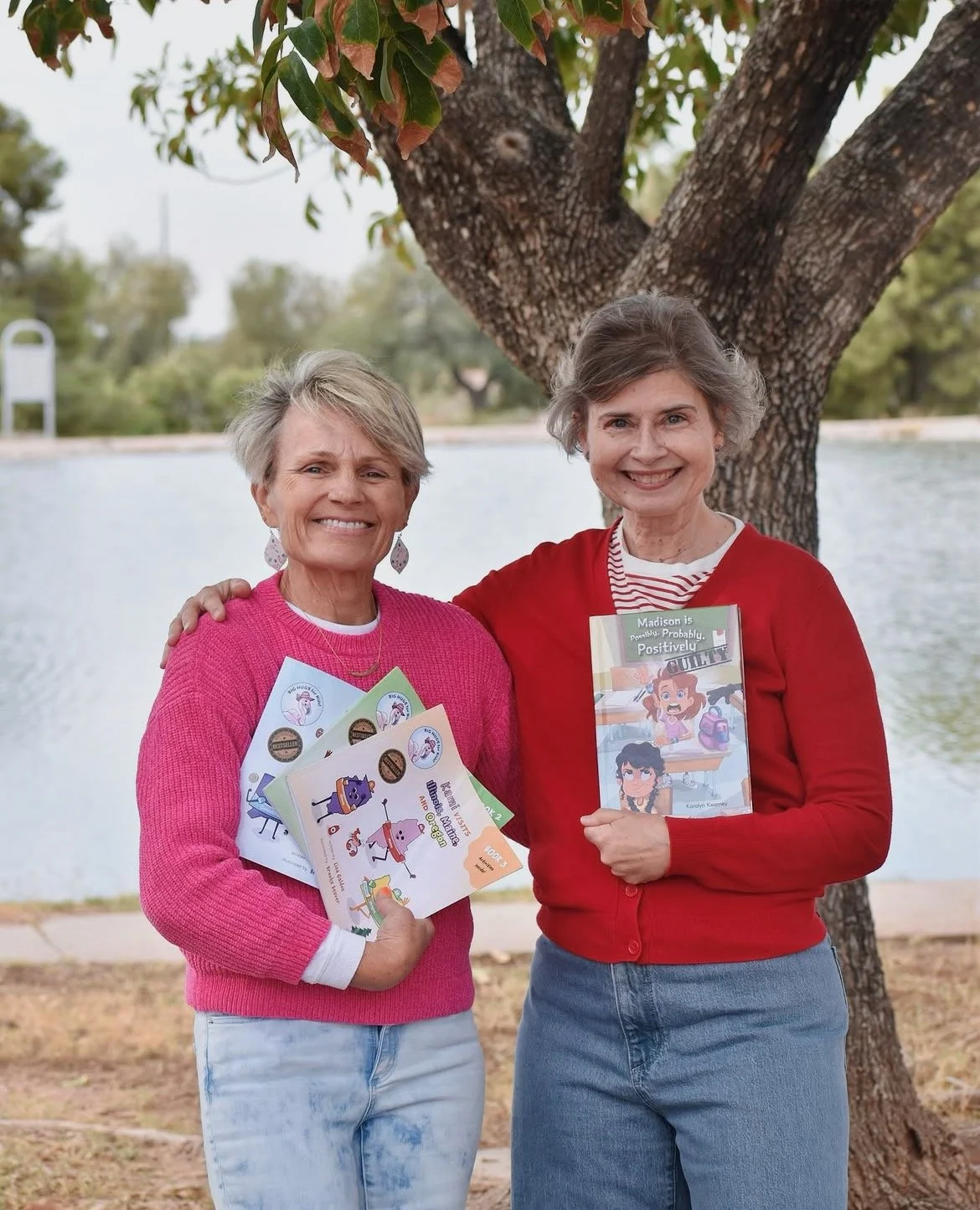 Two women holding children’s books outdoors by a lake, including Madison is Possibly, Probably, Positively Guilty