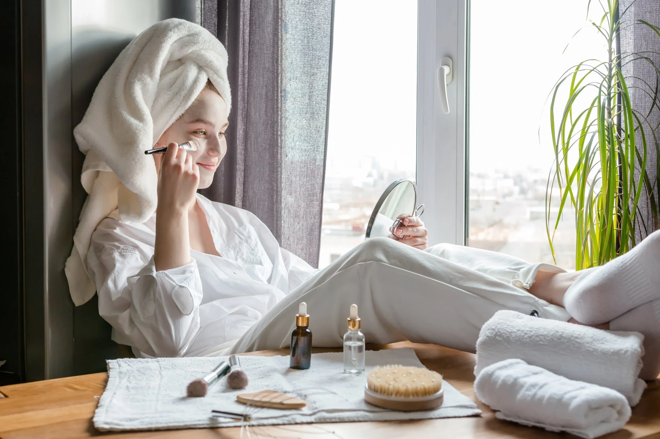 A women in a towel and robe is applying a mask on her face with her feet kicked up on a table.