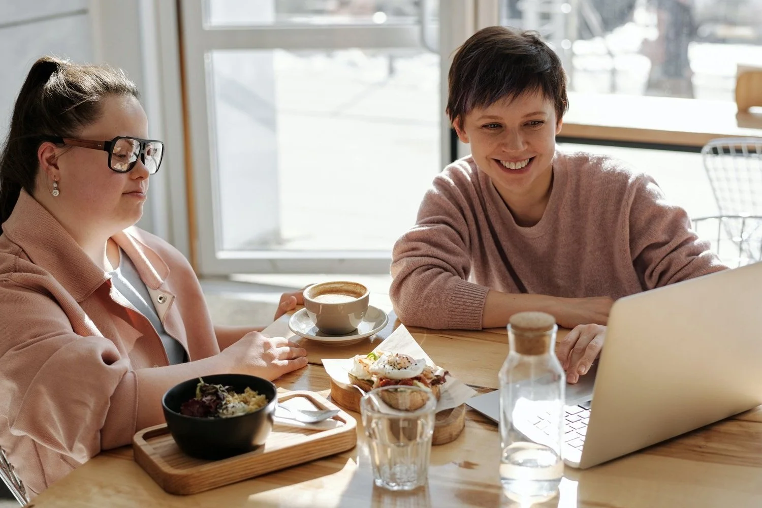two women at a cafe with lunch and coffee, both looking at a laptop screen