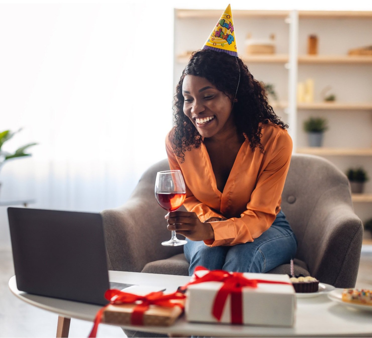 Woman with glass of red wine and a birthday hat on, smiling at a computer screen, and two gift boxes