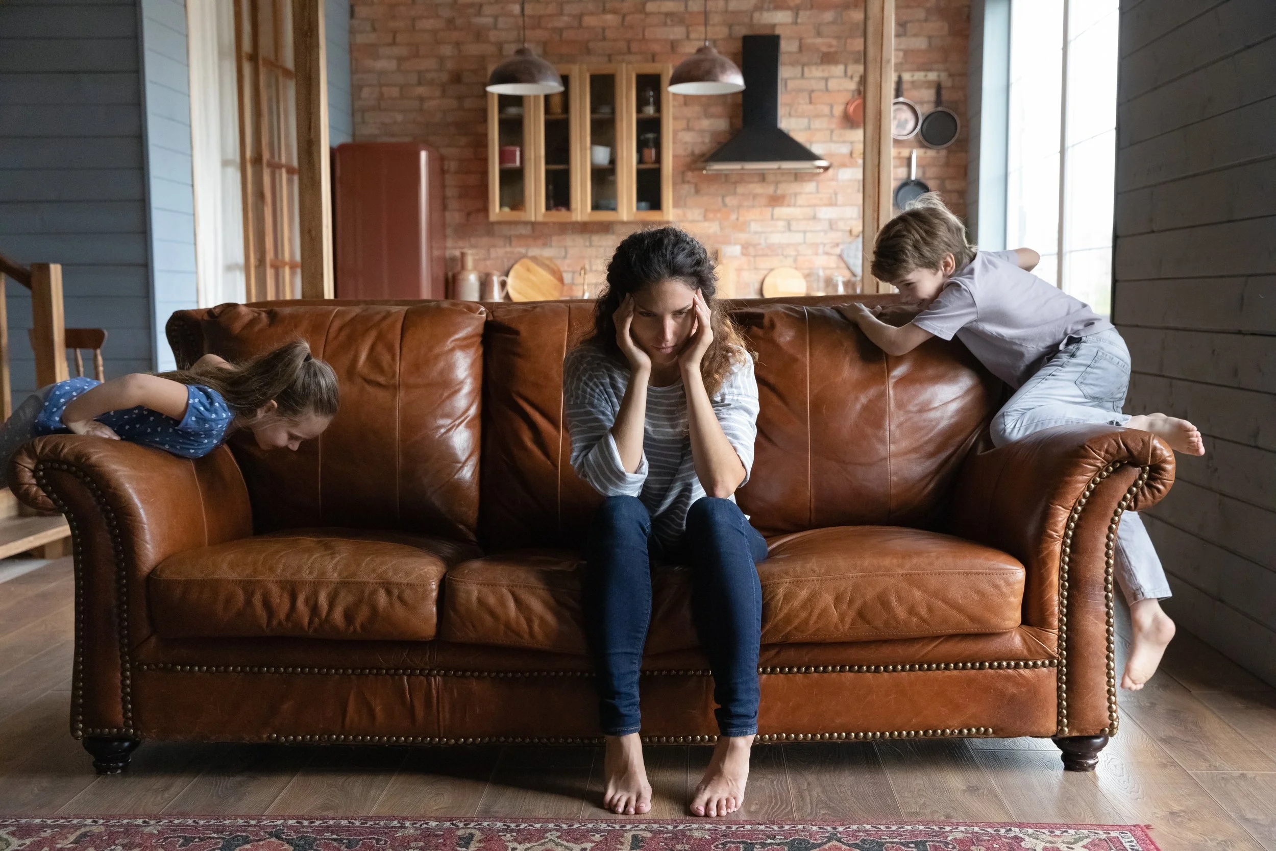 A mother is stressed on a couch with her two kids climbing the couch.
