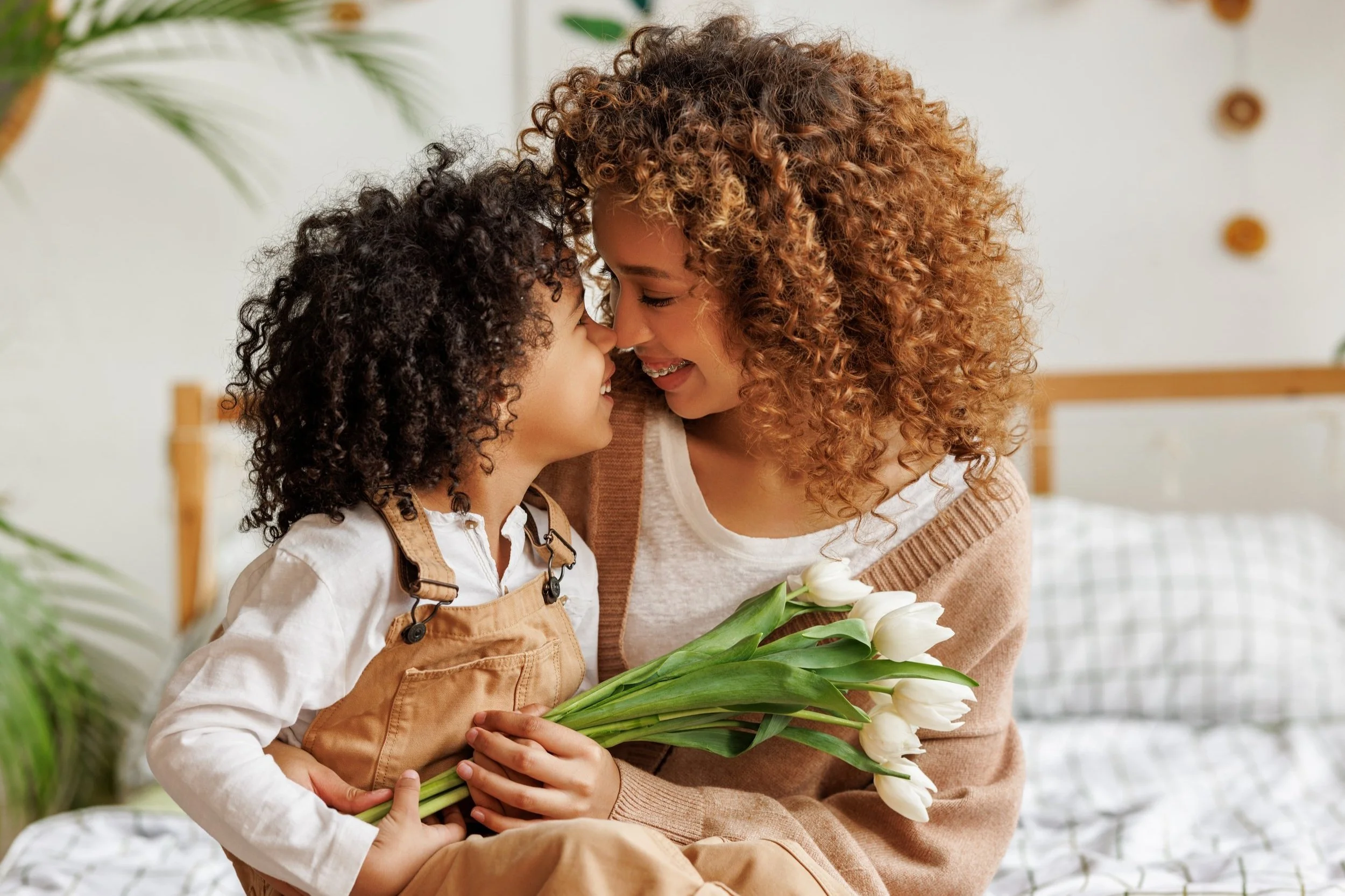 A happy mother on a bed touches her nose to her kids nose while holding white flowers.