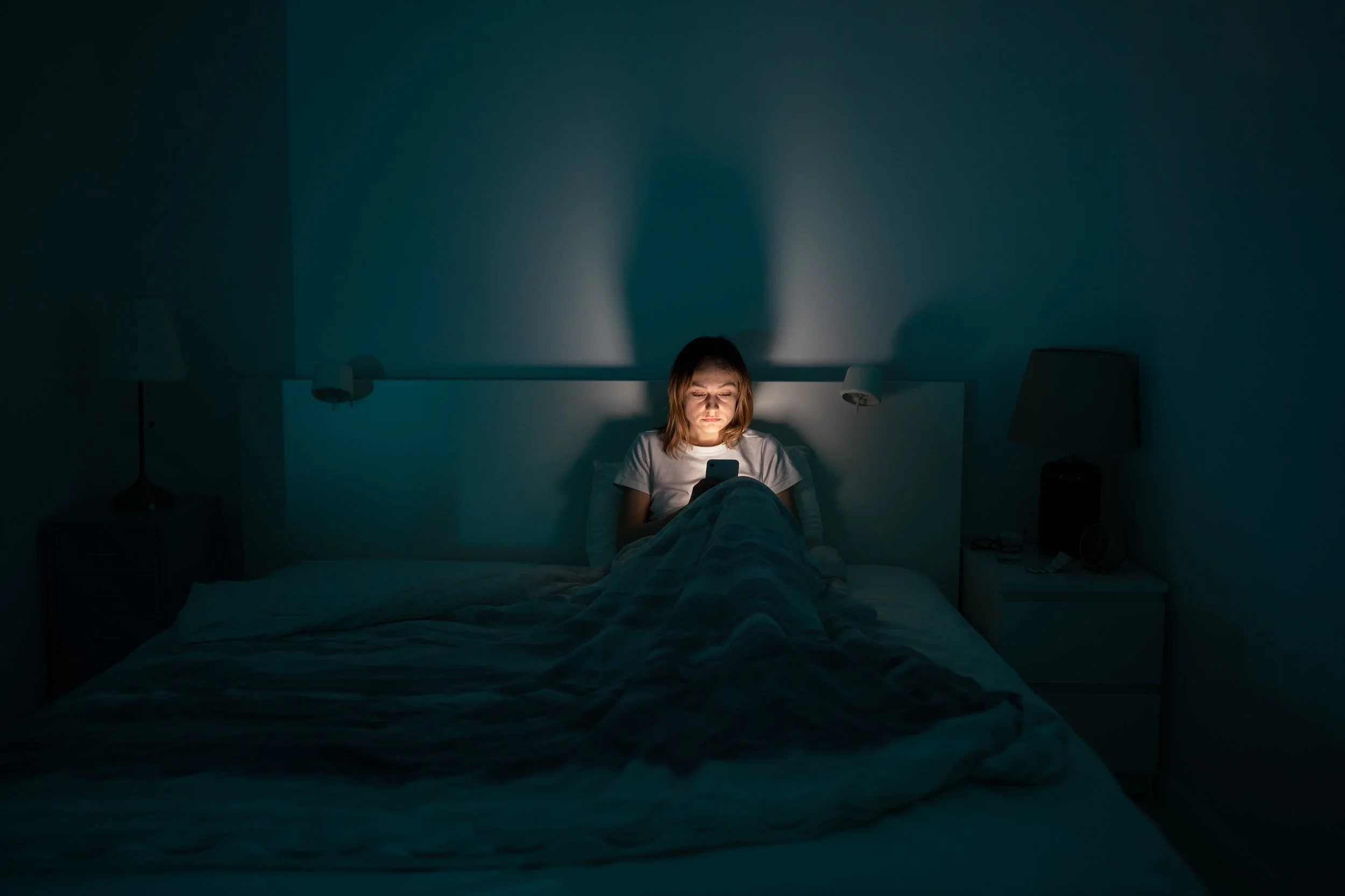 A women in a dark bedroom sits on a bed with a bright cellphone illuminating the room.