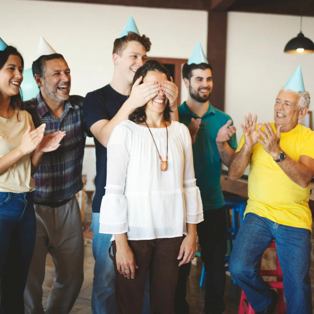 Group of adults with birthday hats on, while one man covers the eyes of a woman to hide a surprise. The other adults are clapping.
