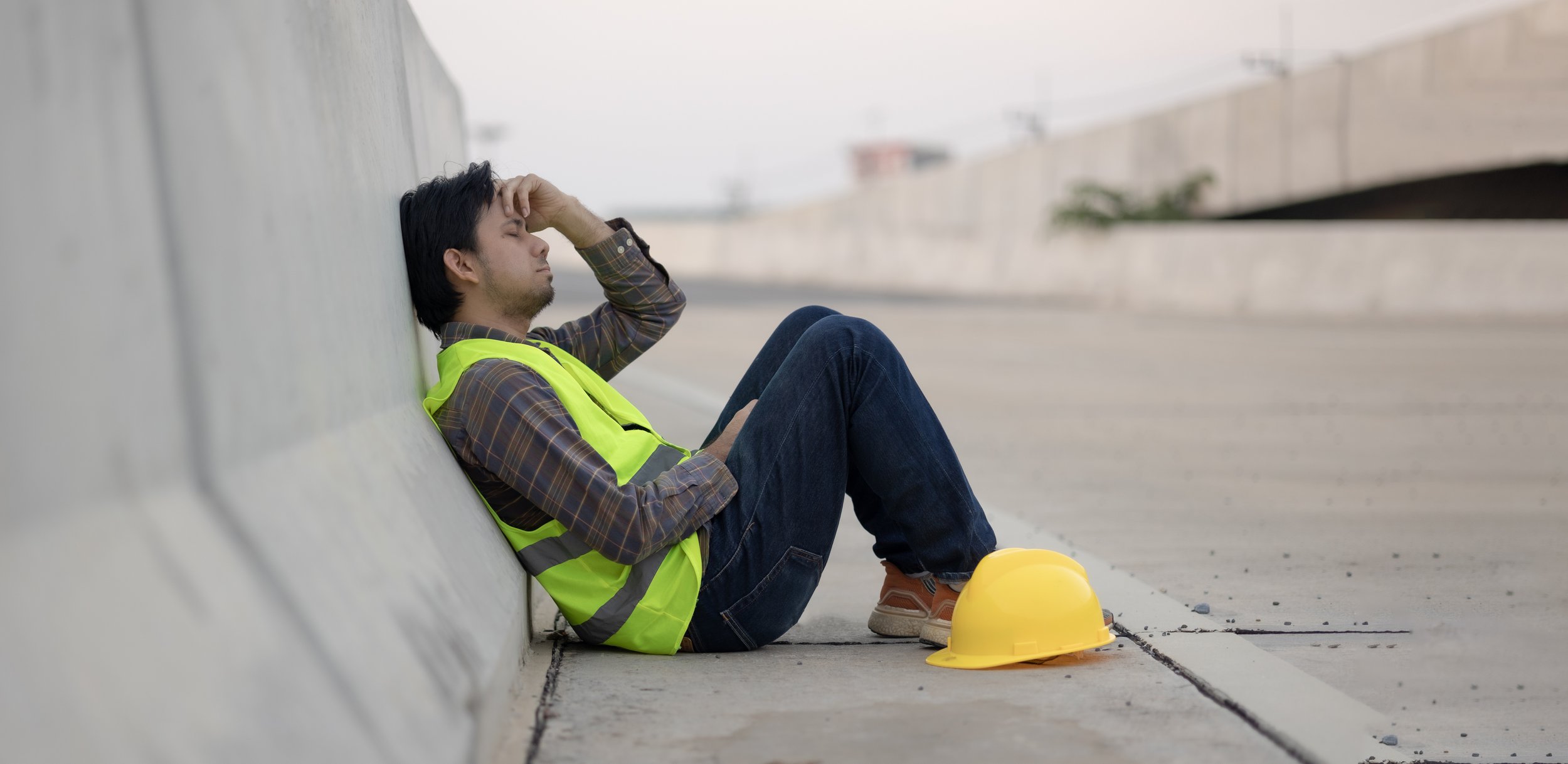 A construction worker is sitting on the ground, back on concrete Jersey barrier. He looks stressed as he holds his head with one hand.