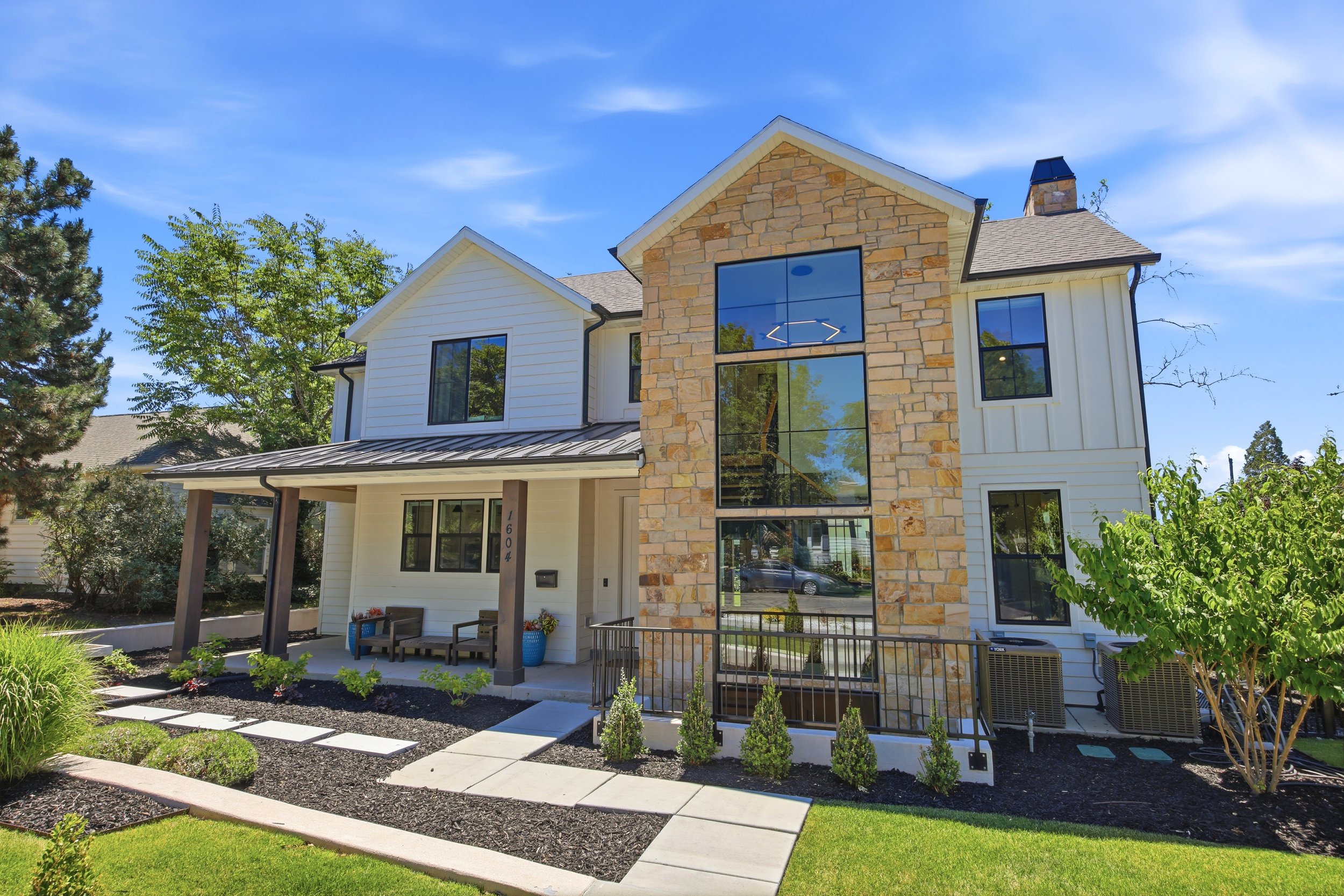 Modern two-story house with white siding and stone accent wall, large windows, a small front porch with benches, well-maintained lawn, garden beds with shrubs, and mature trees in the background under a blue sky.