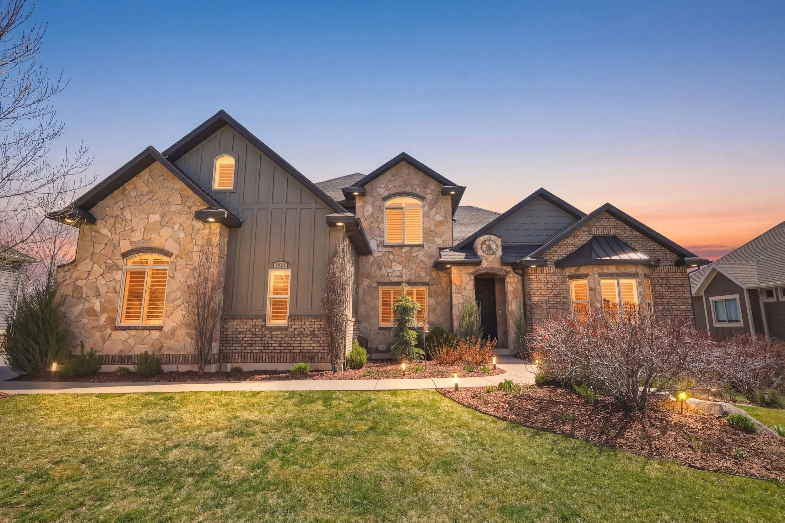 A modern two-story house with stone and siding exterior, illuminated windows, and landscaped front yard at dusk.