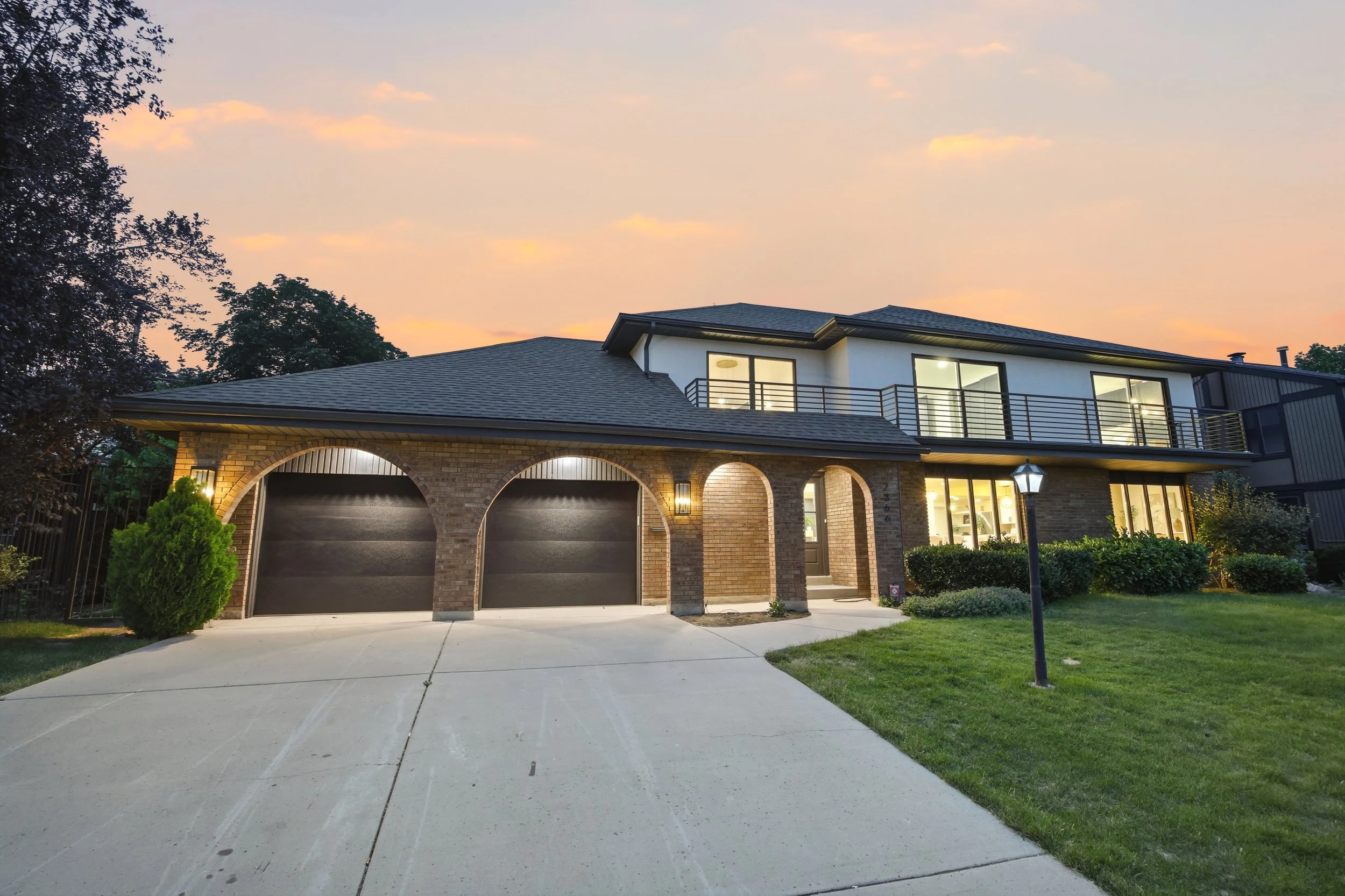 A modern two-story house with a brick and white exterior, large windows, and a balcony on the upper level. The house has a dark roof, garage doors, and a front yard with grass, bushes, and a lamppost, under a sunset sky.