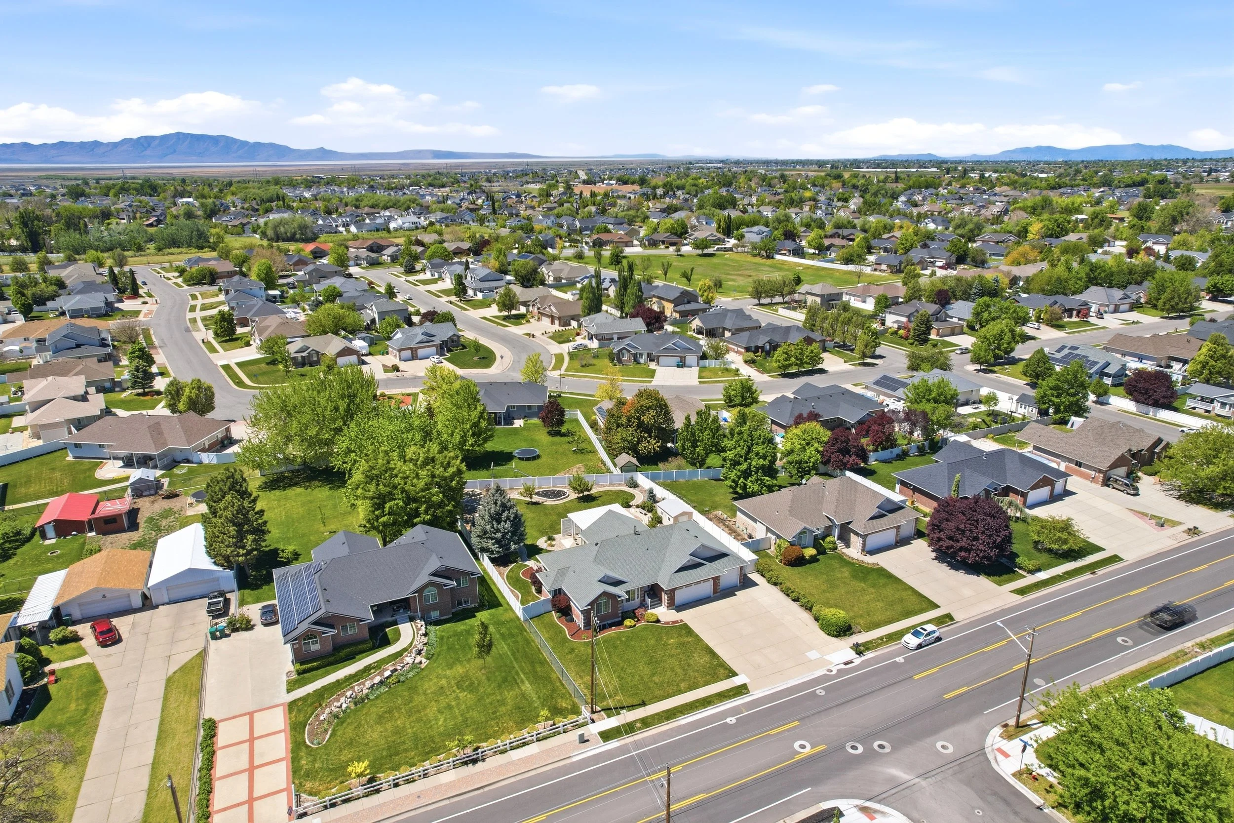 An aerial view of a suburban neighborhood with houses, trees, green lawns, and a parking lot, with mountains and a blue sky in the background.