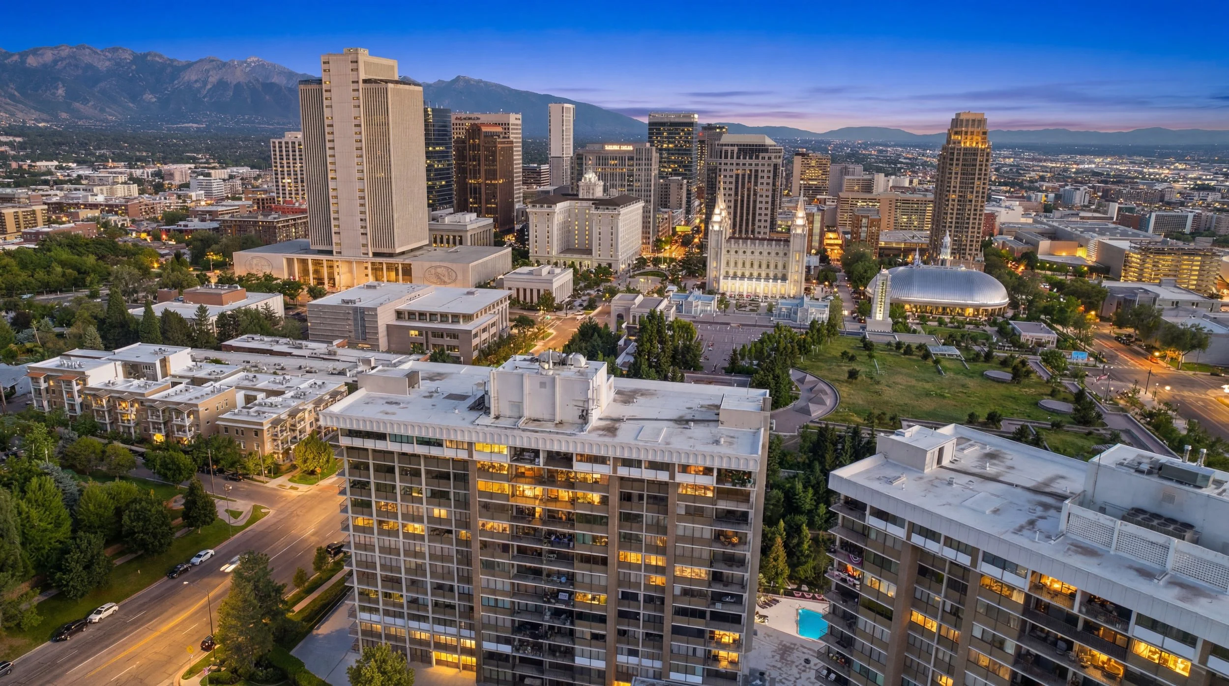 A city skyline at dusk with tall buildings, green parks, and mountains in the background.