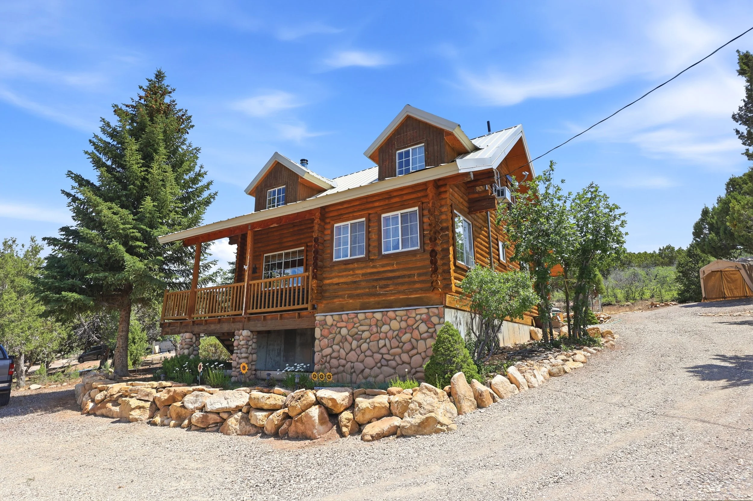 A wooden house with a stone foundation, large windows, and a small front porch, surrounded by trees and a gravel driveway, under a bright blue sky with a few clouds.
