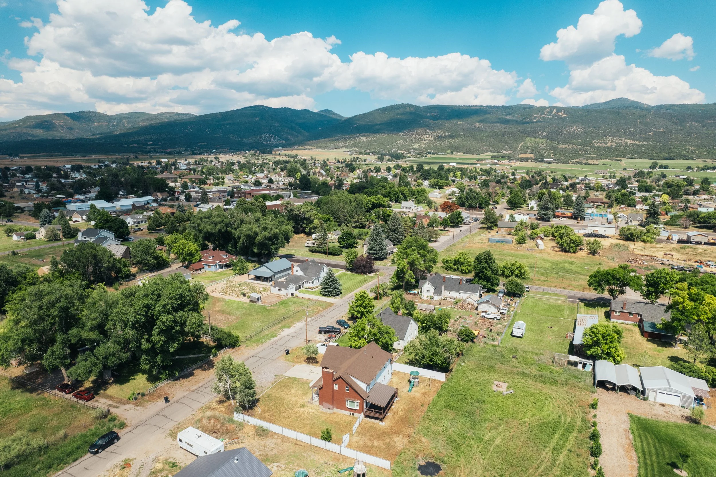 Aerial view of a small town with houses, streets, trees, and open green spaces, with mountains in the background under a partly cloudy sky.