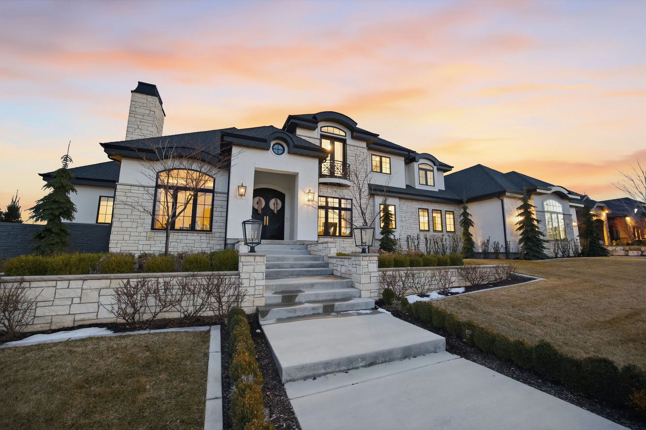 Front view of large modern house with stone and white exterior, black roof, large windows, and a staircase leading to the front door, during sunset.