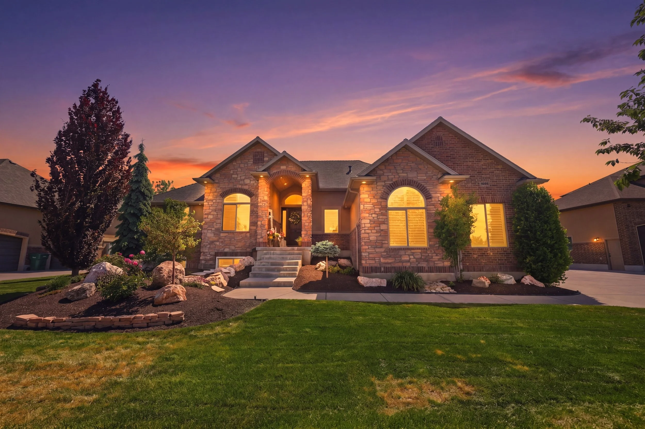 A modern brick and stone house with a well-maintained front yard at sunset, illuminated windows, and landscaped garden.