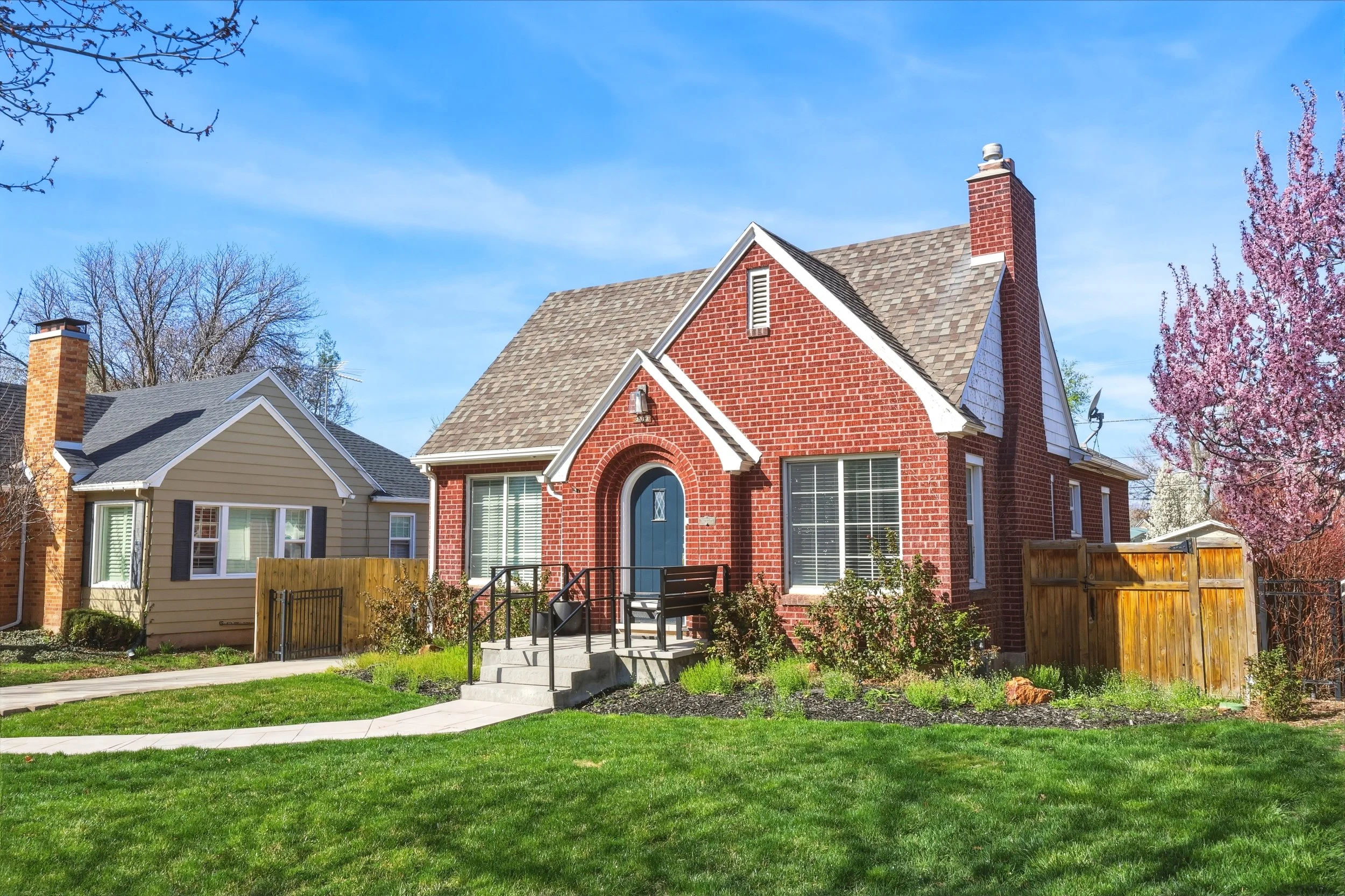 Quaint red brick bungalow with an arched blue front door, mature landscaping, green lawn, and blooming spring trees.