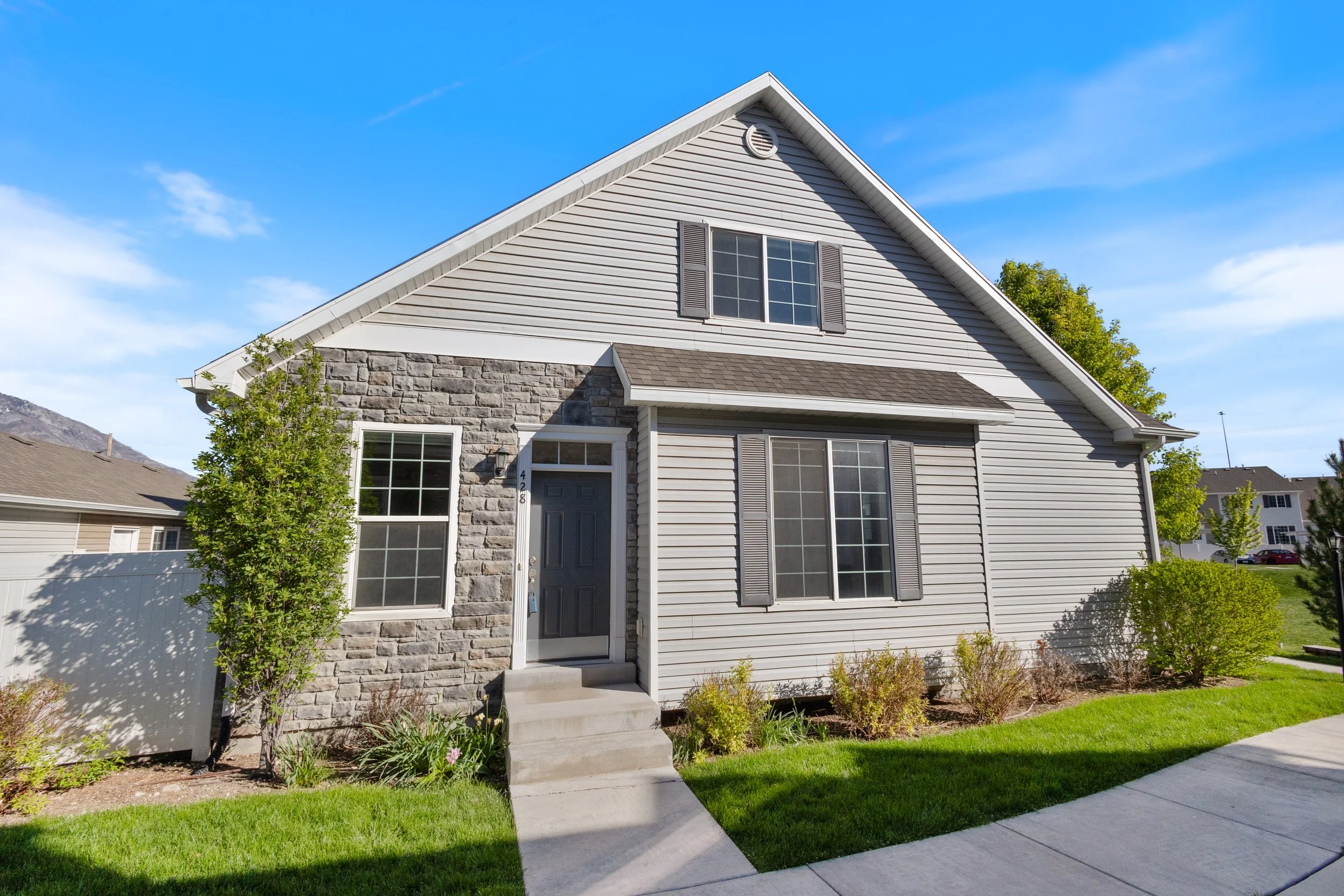 Front view of a two-story house with grey siding, stone accents, and a gabled roof against a blue sky.