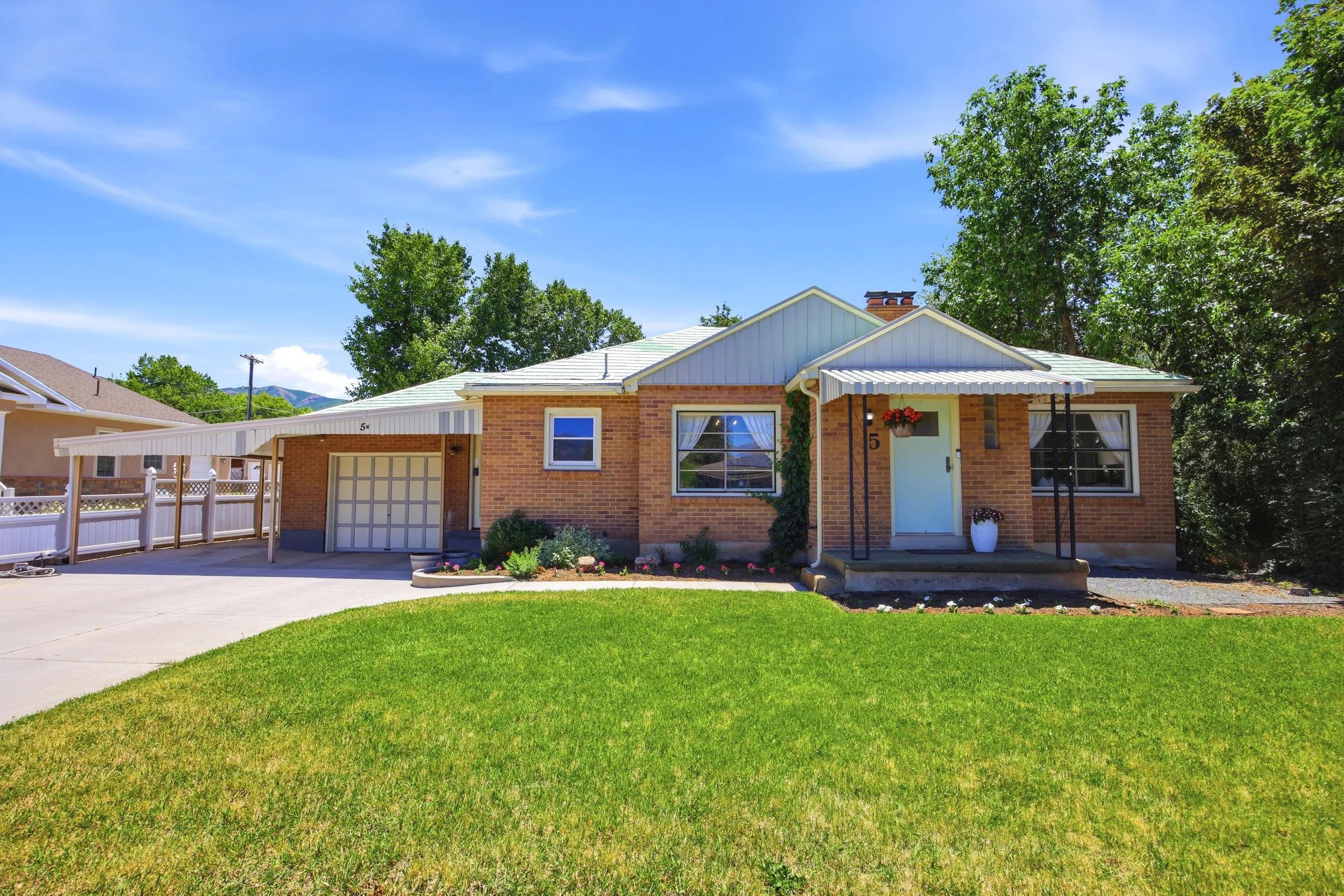 A suburban house with brick exterior, white trim, and a metal roof, surrounded by green trees and a well-maintained lawn, under a clear blue sky.