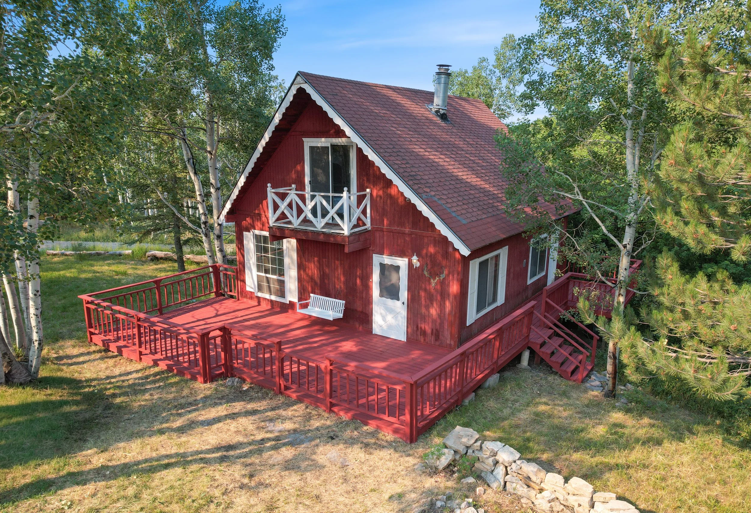 Red wooden house with white trim, a small balcony, and a deck, surrounded by green trees and blue sky.