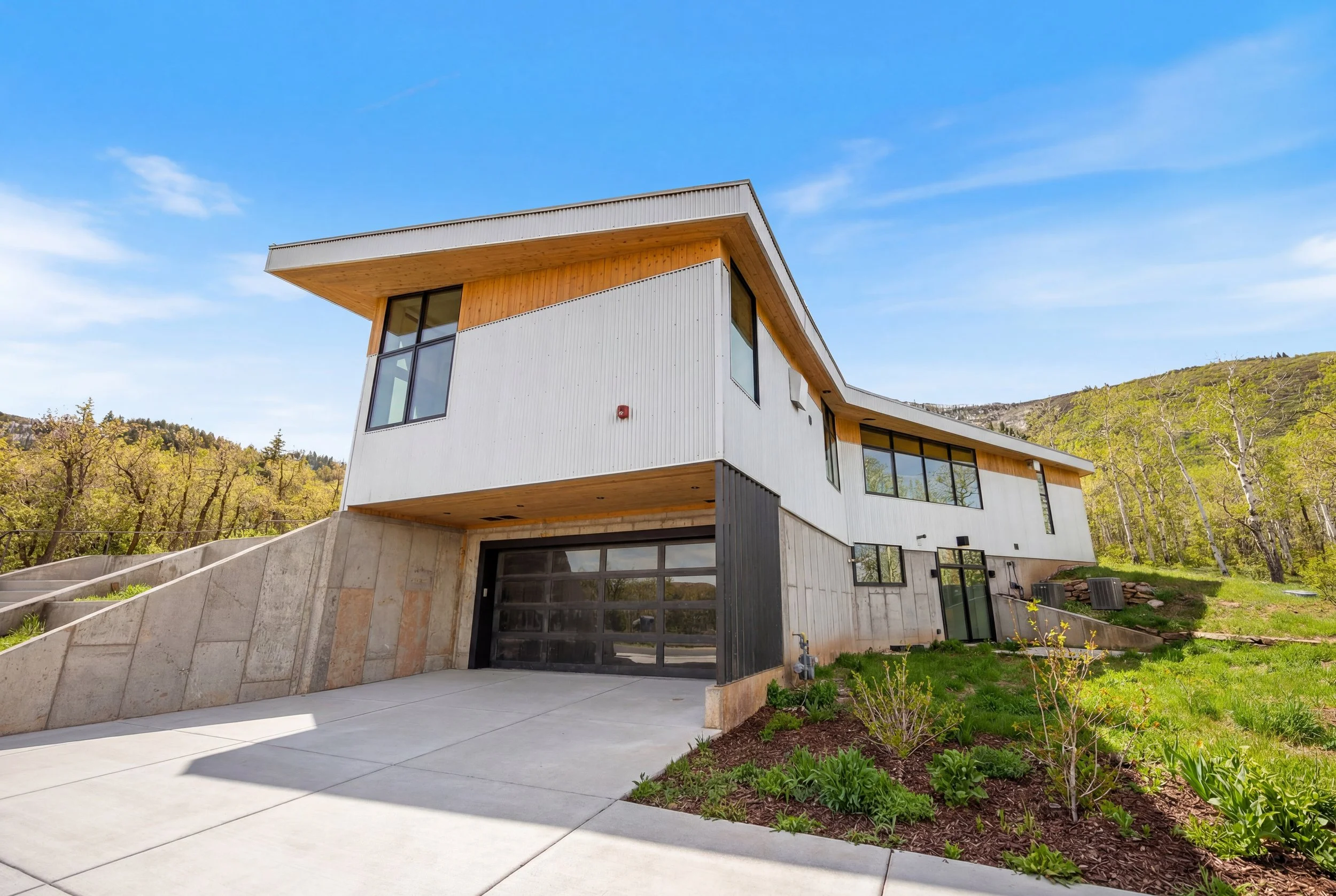 Modern two-story house with white siding, large windows, and wooden accents, set against a backdrop of trees and hills under a blue sky.
