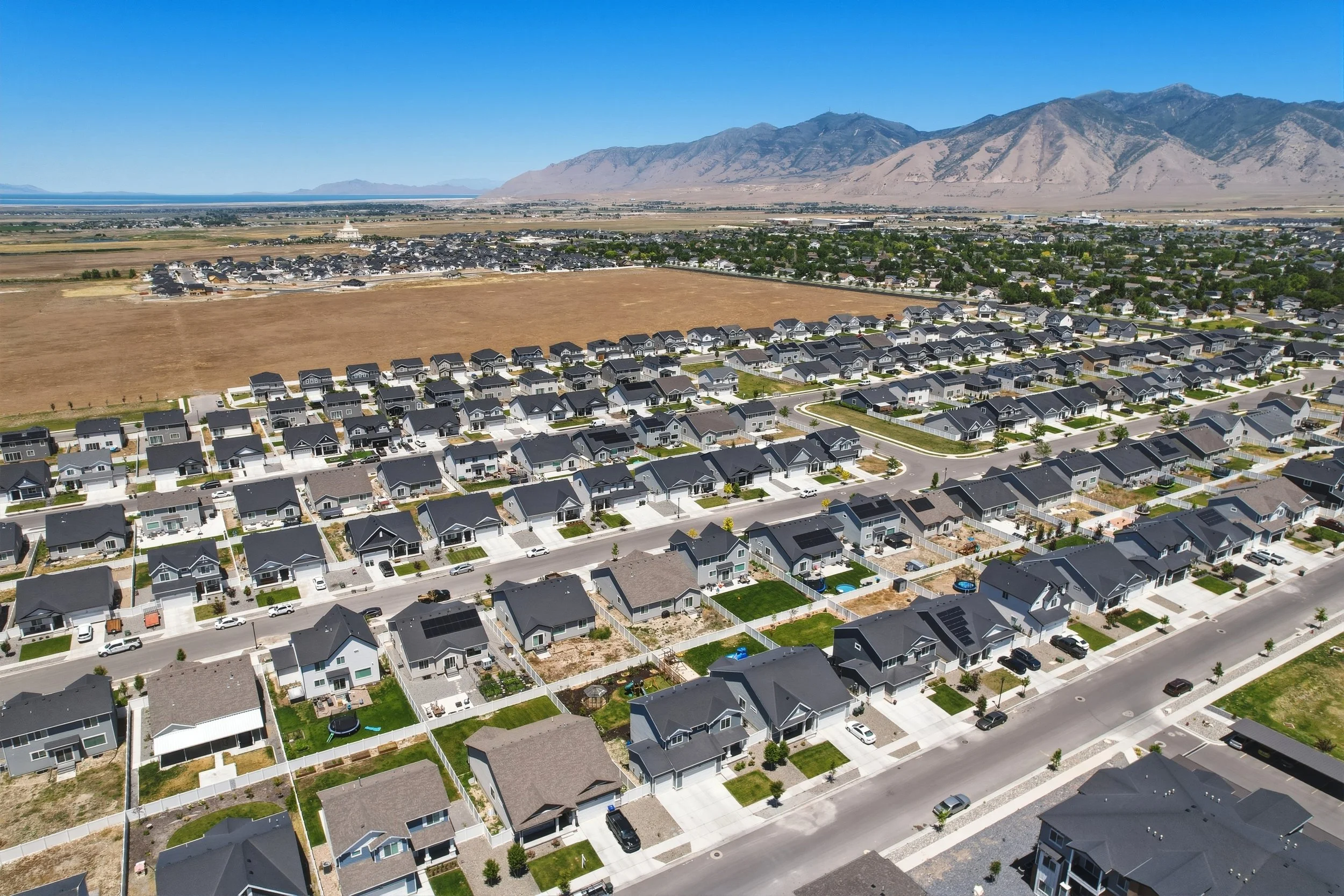 Aerial view of a residential neighborhood with houses, streets, yards, and a large open field. Mountains are visible in the background under a clear blue sky.