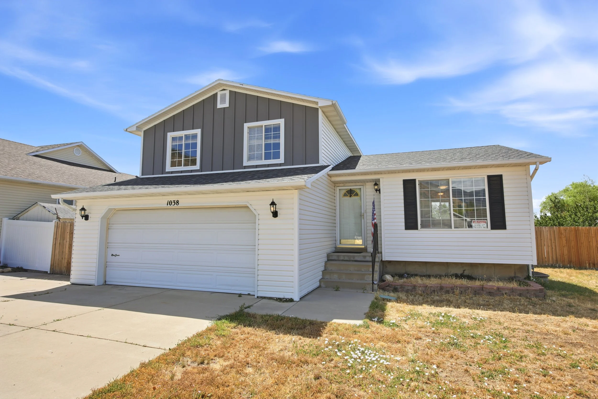 Front view of a two-story suburban house with a white garage door, gray siding on the upper level, white siding on the lower level, and a small porch with steps. The yard has patchy grass and a concrete driveway under a clear blue sky.