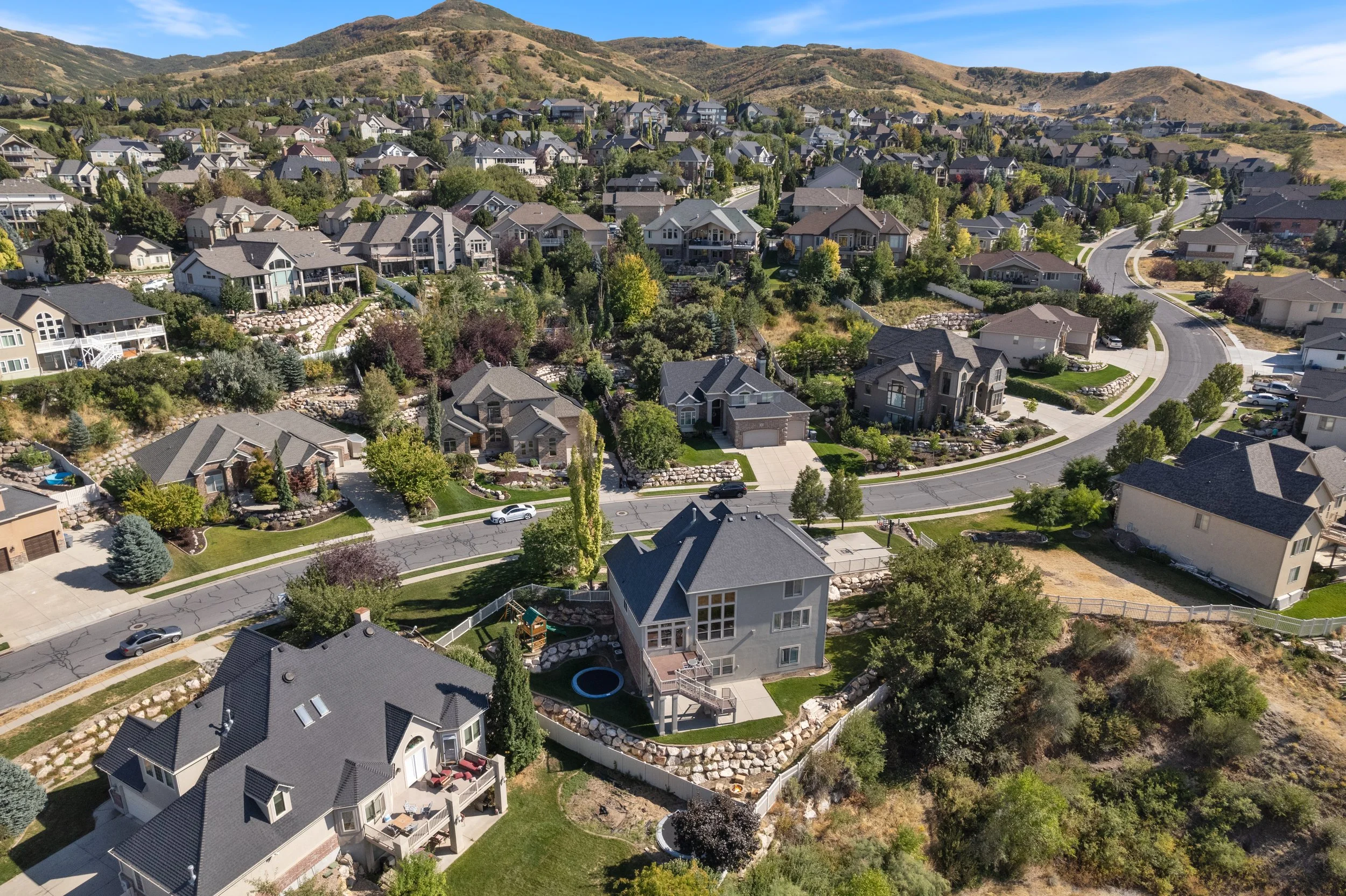 Aerial view of a residential neighborhood with houses, trees, and winding roads, set against a backdrop of rolling hills under a clear blue sky.