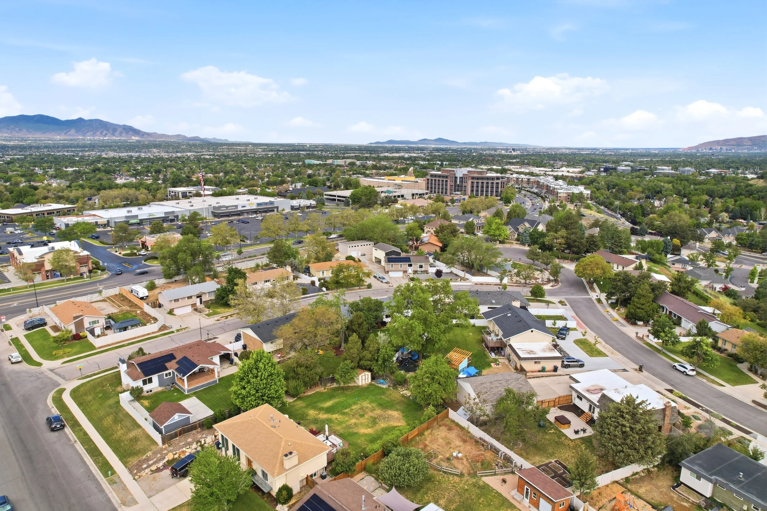 An aerial view of a suburban neighborhood with houses, trees, and streets, with a cityscape and mountains in the distance under a partly cloudy sky.