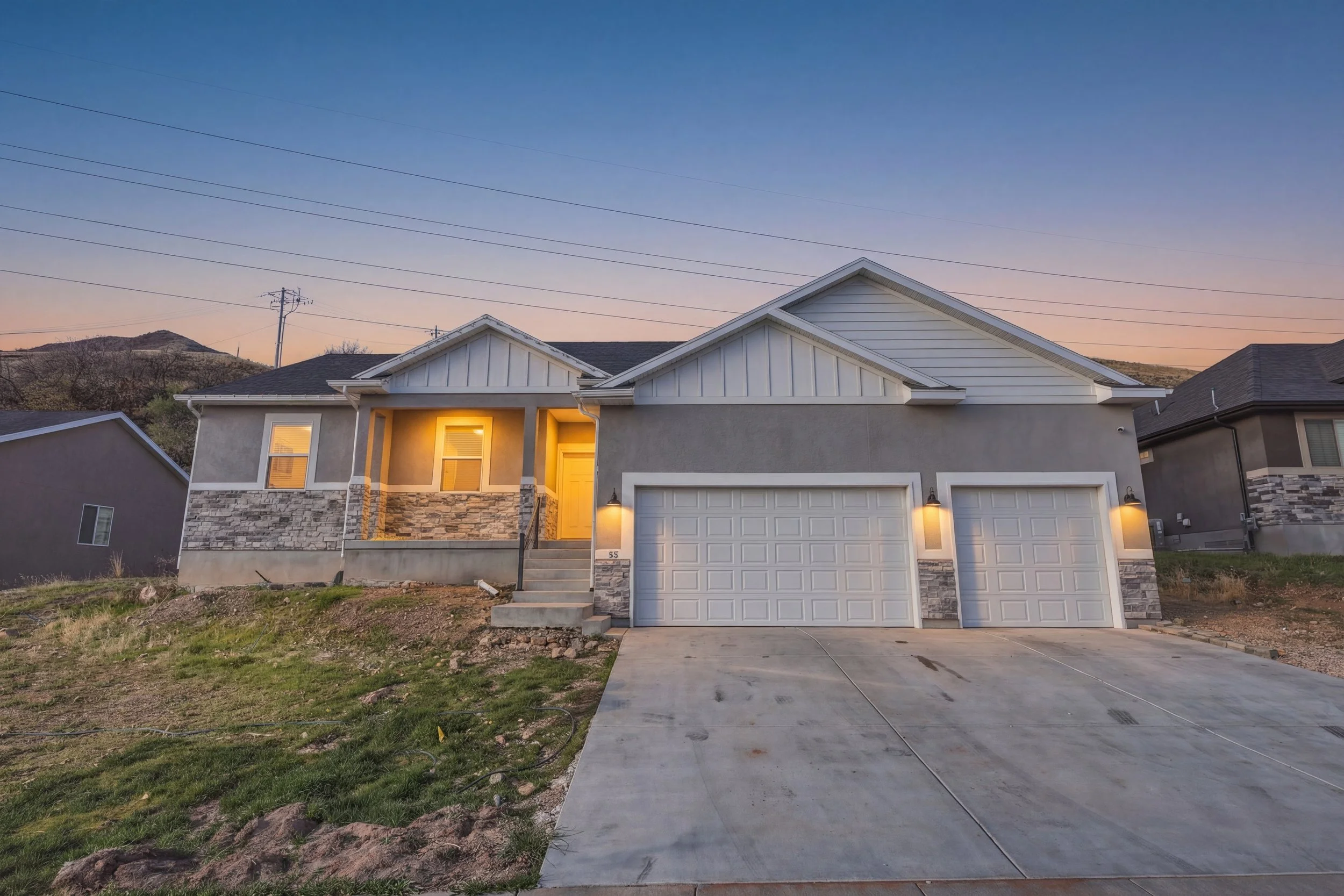 A modern two-story house with a stone and stucco exterior, illuminated porch lights, double garage doors, and a concrete driveway, set against a backdrop of hills and a clear twilight sky.